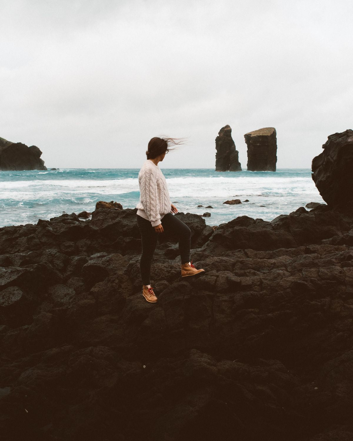 Woman wearing LOBO Raw Boots on black lava rocks at Mosteiros Beach, with sea stacks in the background on a stormy day.