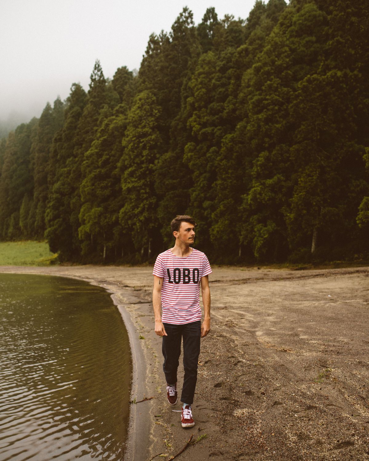 Young man walking along the shore of Lagoa de São Brás, Azores, wearing a red and white striped t-shirt with the LOBO logo. In the background, a dense Japanese cedar forest under a misty sky.