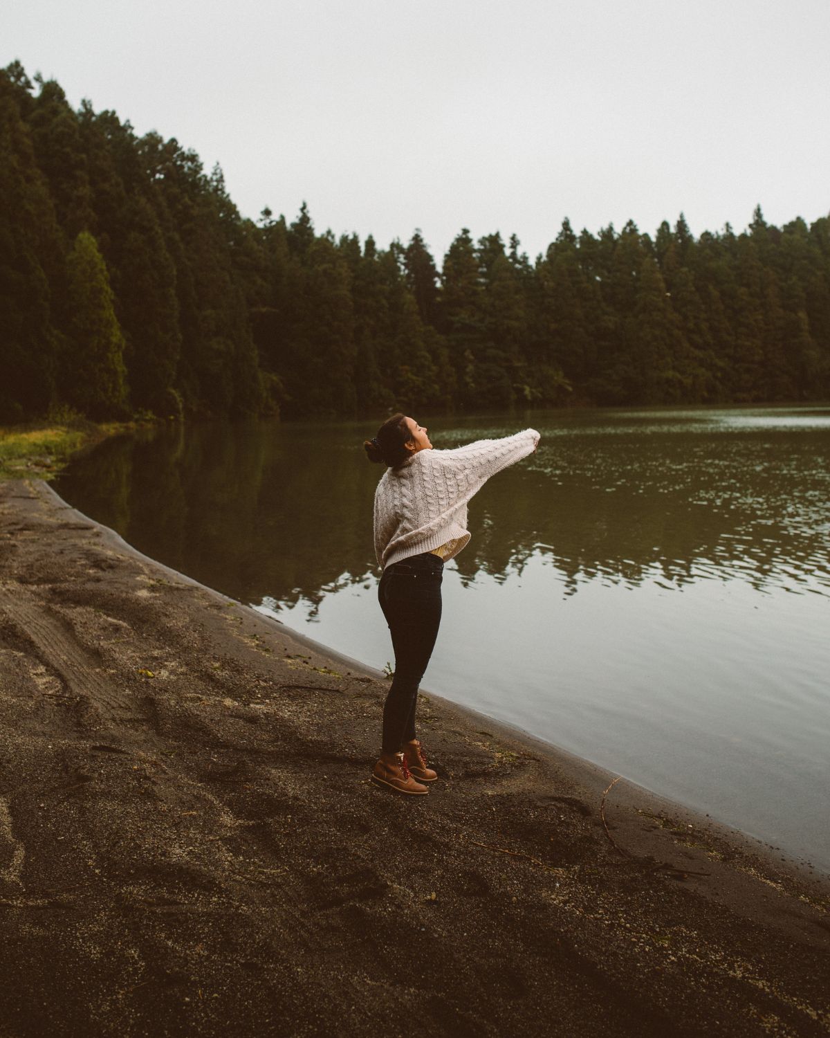 Woman wearing a knit sweater and LOBO Raw boots looking at Lagoa de São Brás surrounded by a cedar forest.