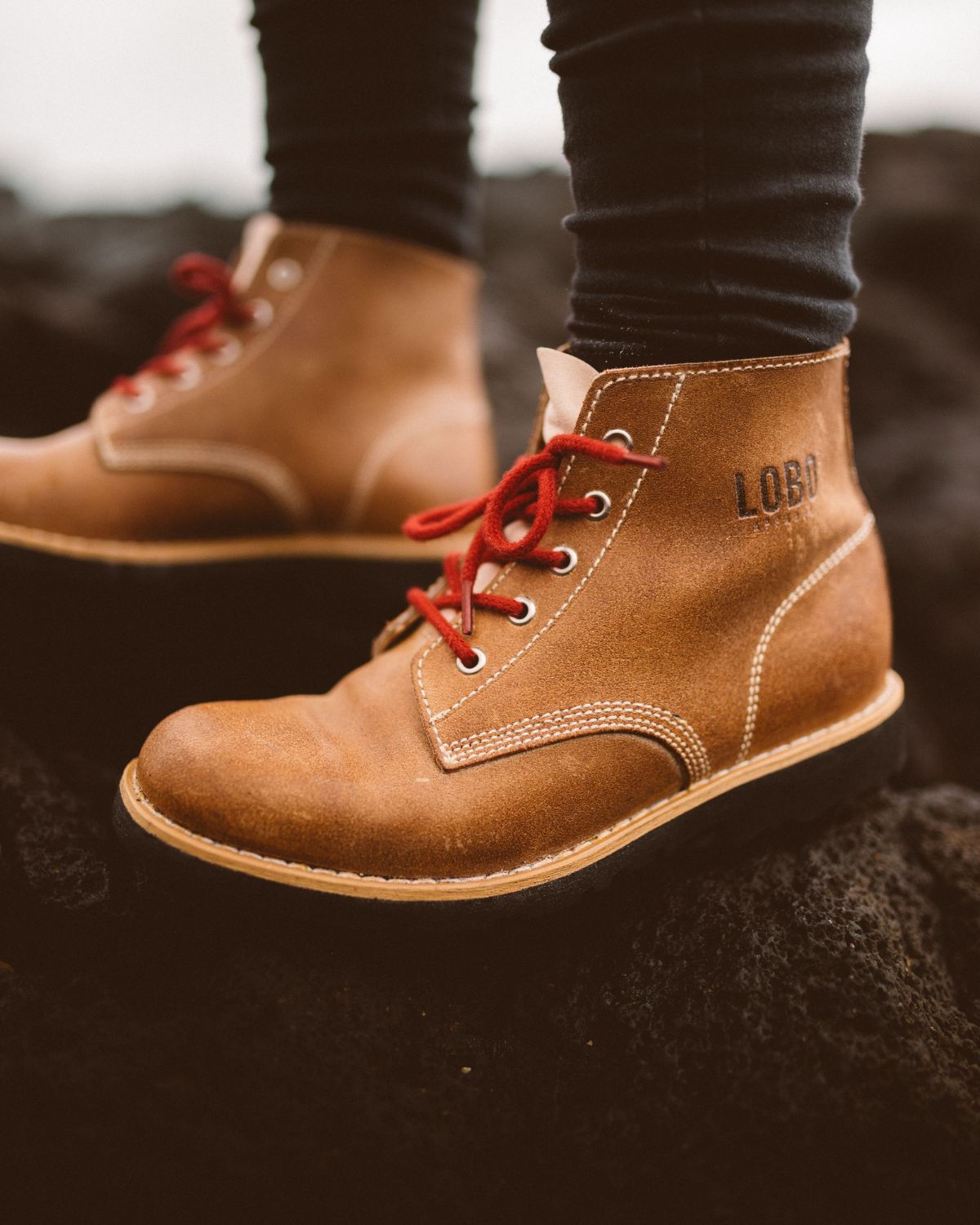 Close-up of LOBO camel leather boots with red laces and embossed logo, standing on volcanic rock.