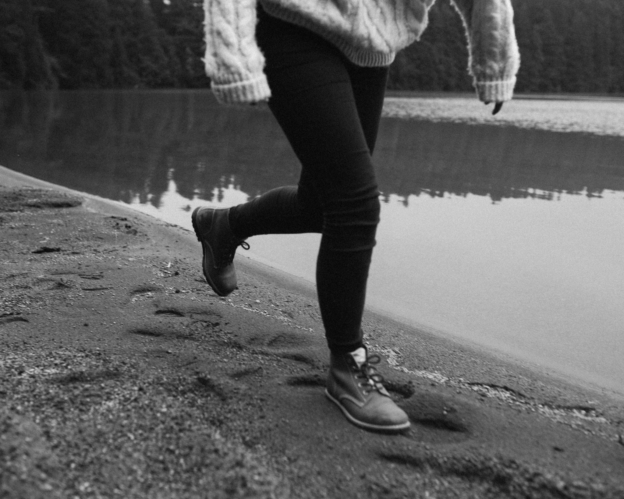 Artistic black and white photograph of a person walking on volcanic sand wearing Lobo Raw Boots. The focus is on the leather texture of the boots and the footprint left on the lagoon's shore, conveying durability and adventure.
