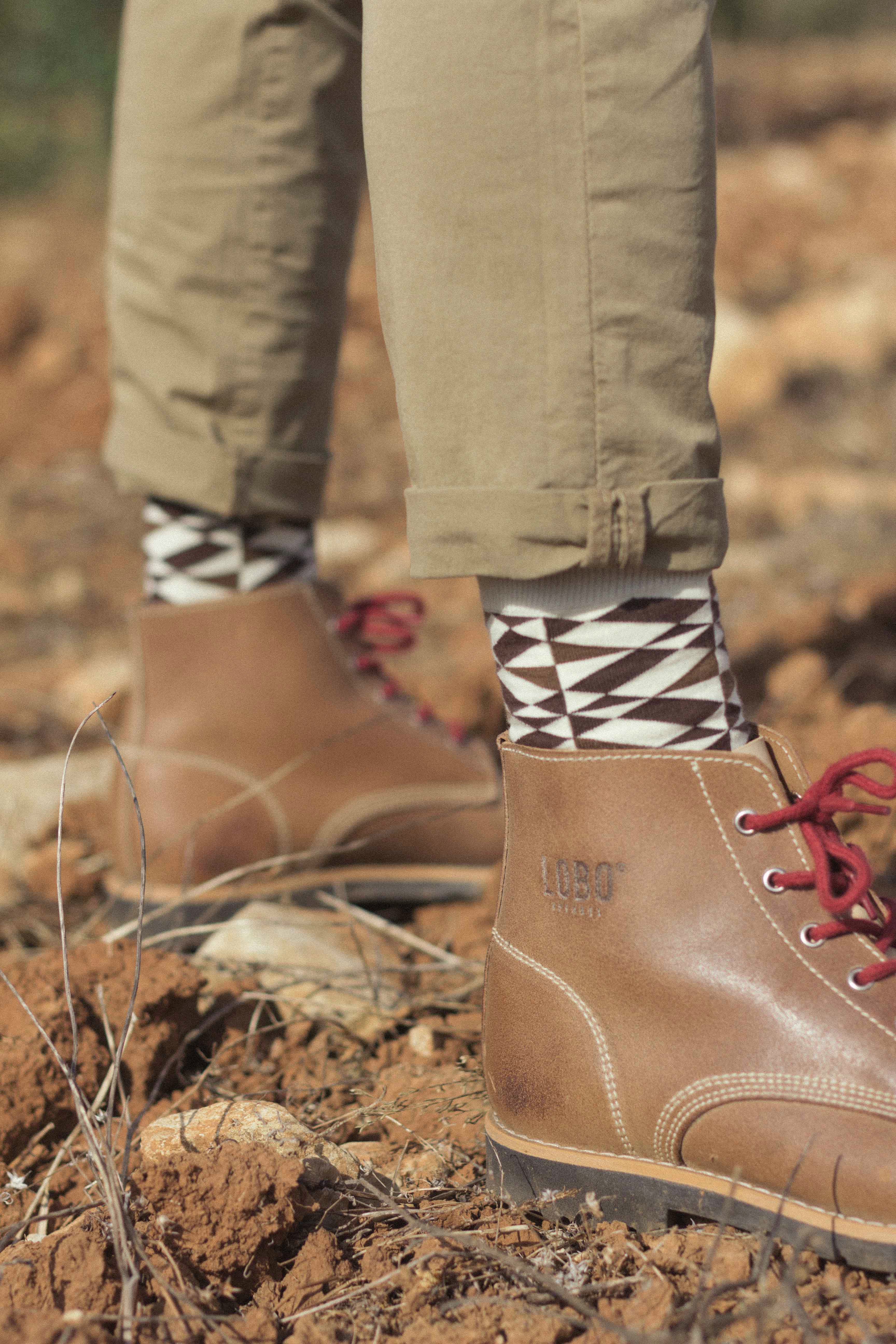 Close-up of Lobo Raw Boots in brown leather with red laces, paired with geometric pattern socks on a nature trail.