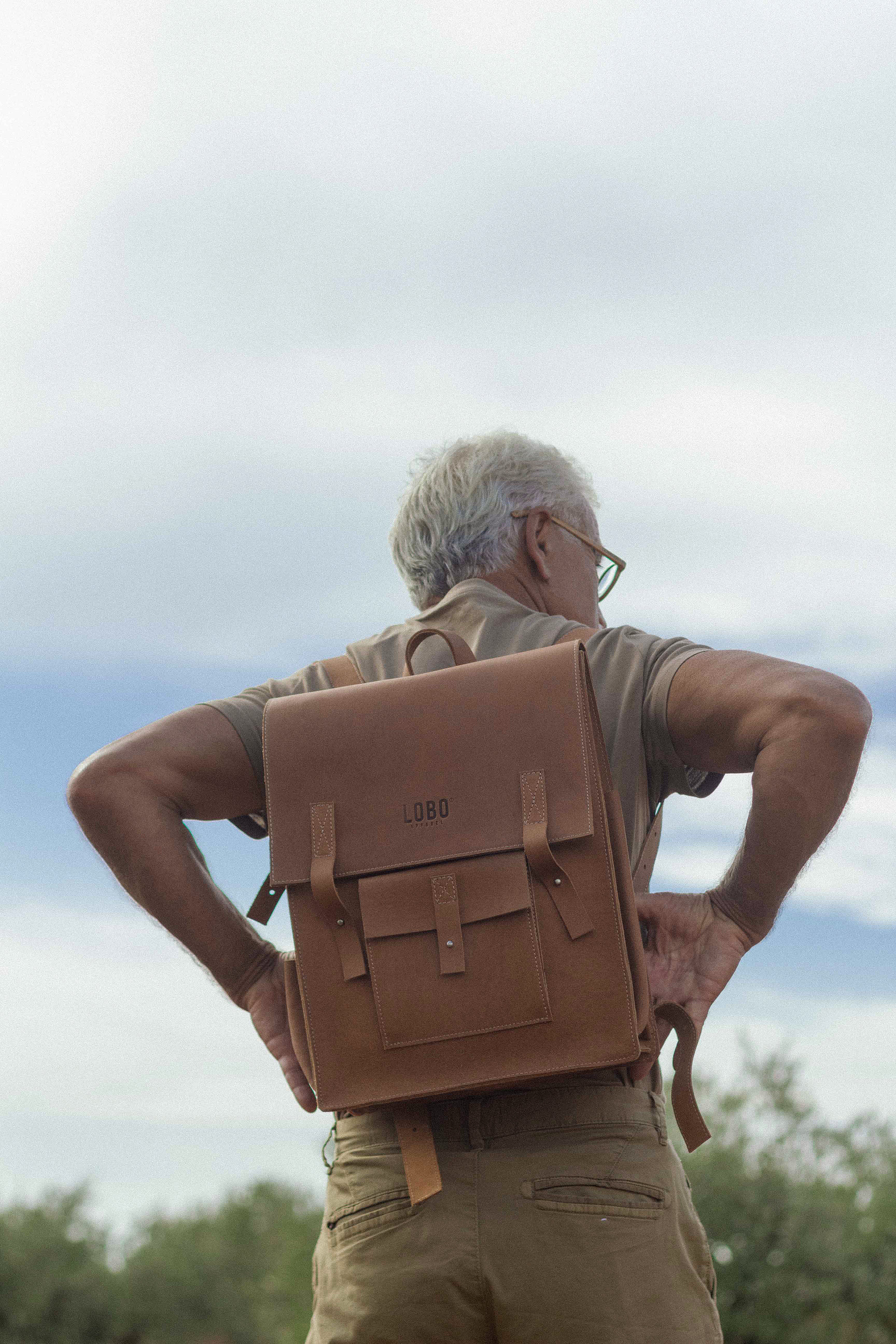 Back view of a man hiking outdoors wearing a brown leather Lobo backpack, evoking exploration and freedom in nature.