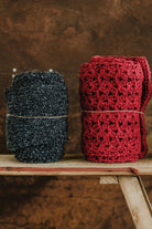 Two handmade Re-Born scarves — one in heather grey and another in burgundy crochet — crafted from repurposed materials, displayed on a wooden bench.