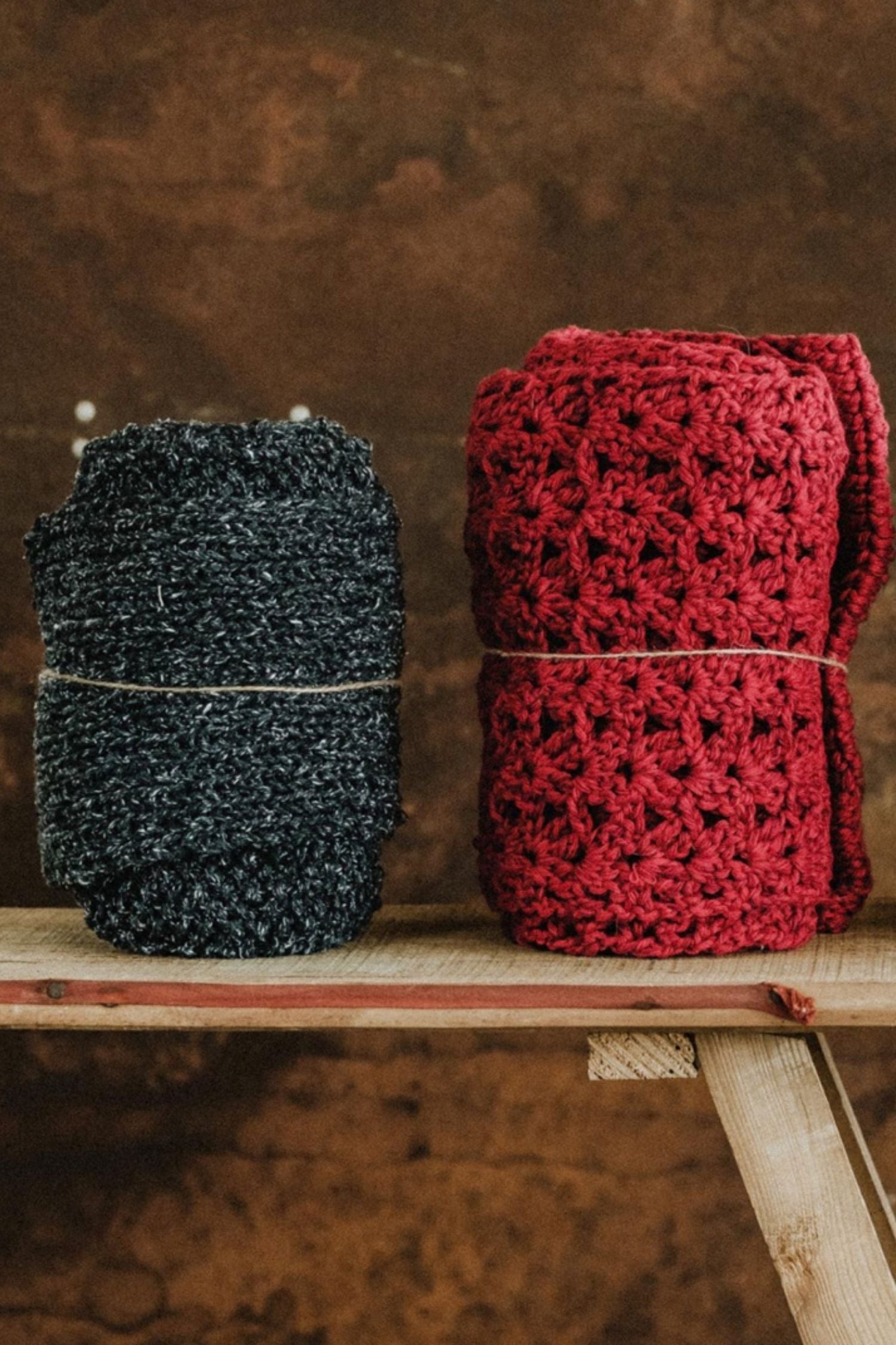 Two handmade Re-Born scarves — one in heather grey and another in burgundy crochet — crafted from repurposed materials, displayed on a wooden bench.