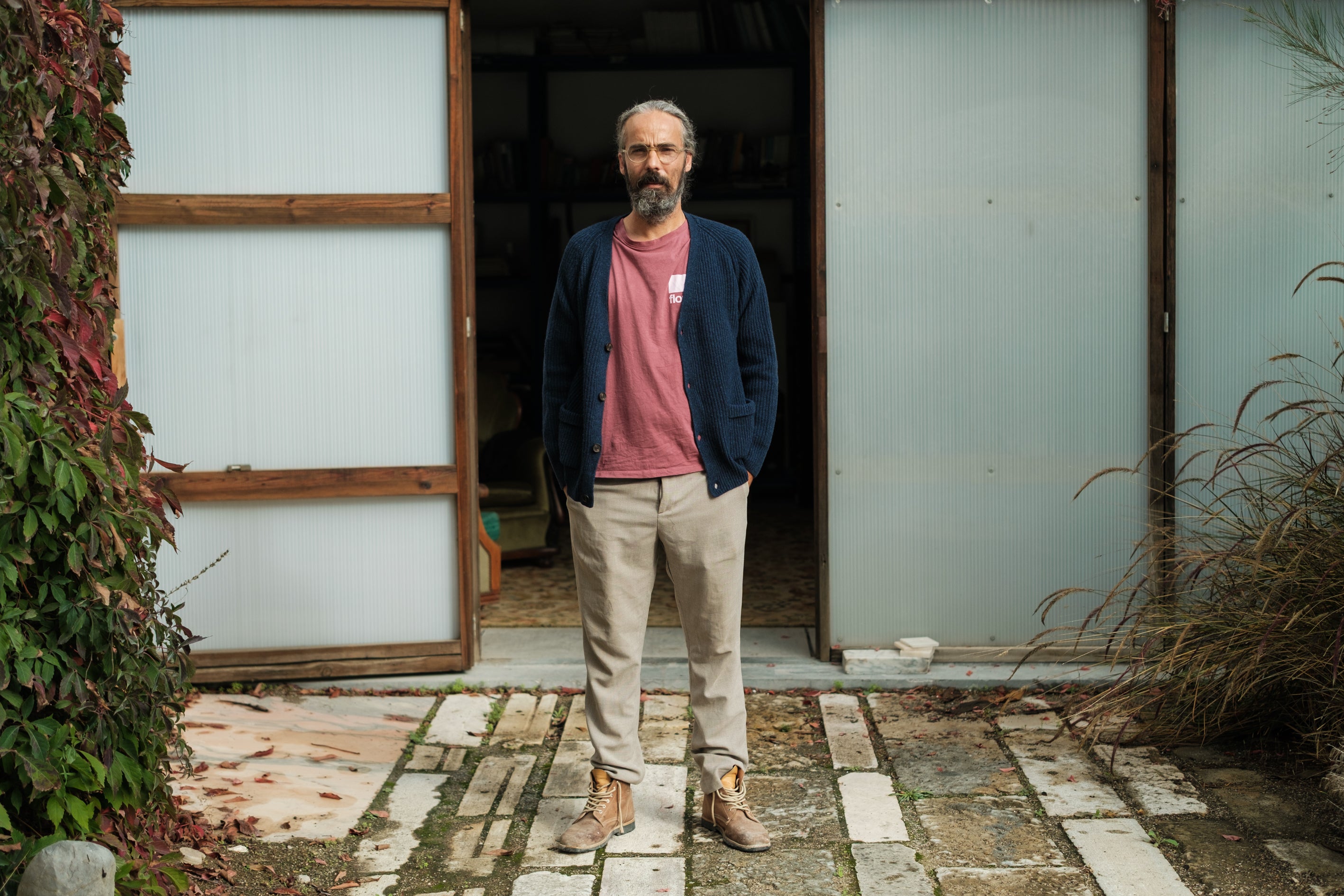 Full-length portrait of architect Carlos Flores standing in front of his studio. He is wearing a blue cardigan, beige trousers, and his brown Lobo Raw Boots with a natural worn patina. The outdoor setting with stone flooring and greenery reinforces the authentic and durable aesthetic of the Lobo Apparel brand.