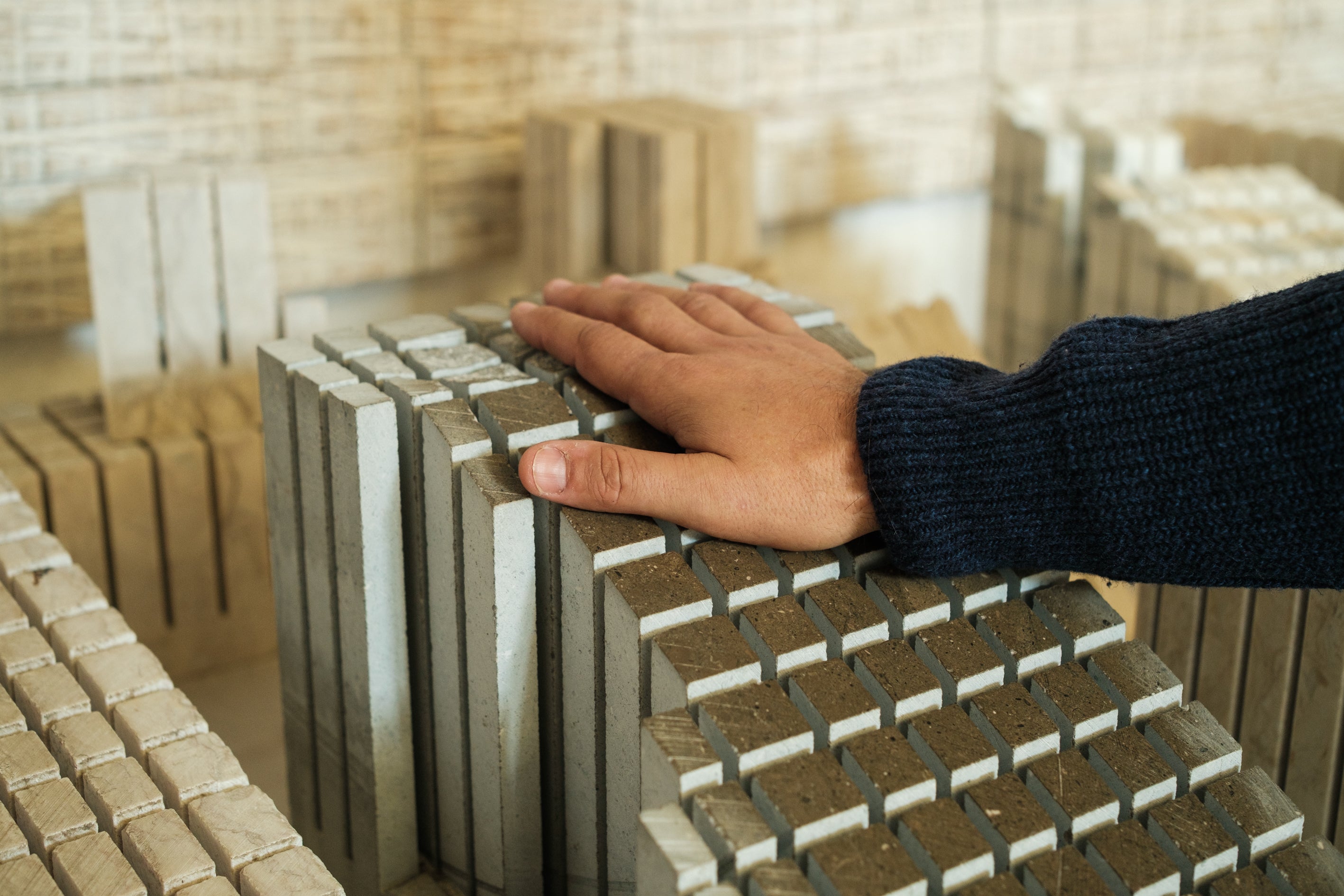Close-up of Carlos Flores' hand, wearing a dark blue knit sweater, touching structured stone blocks in a workshop. The image highlights the texture of the material and the tactile connection between the architect and the raw stone, evoking durability and his "son of stone" philosophy.
