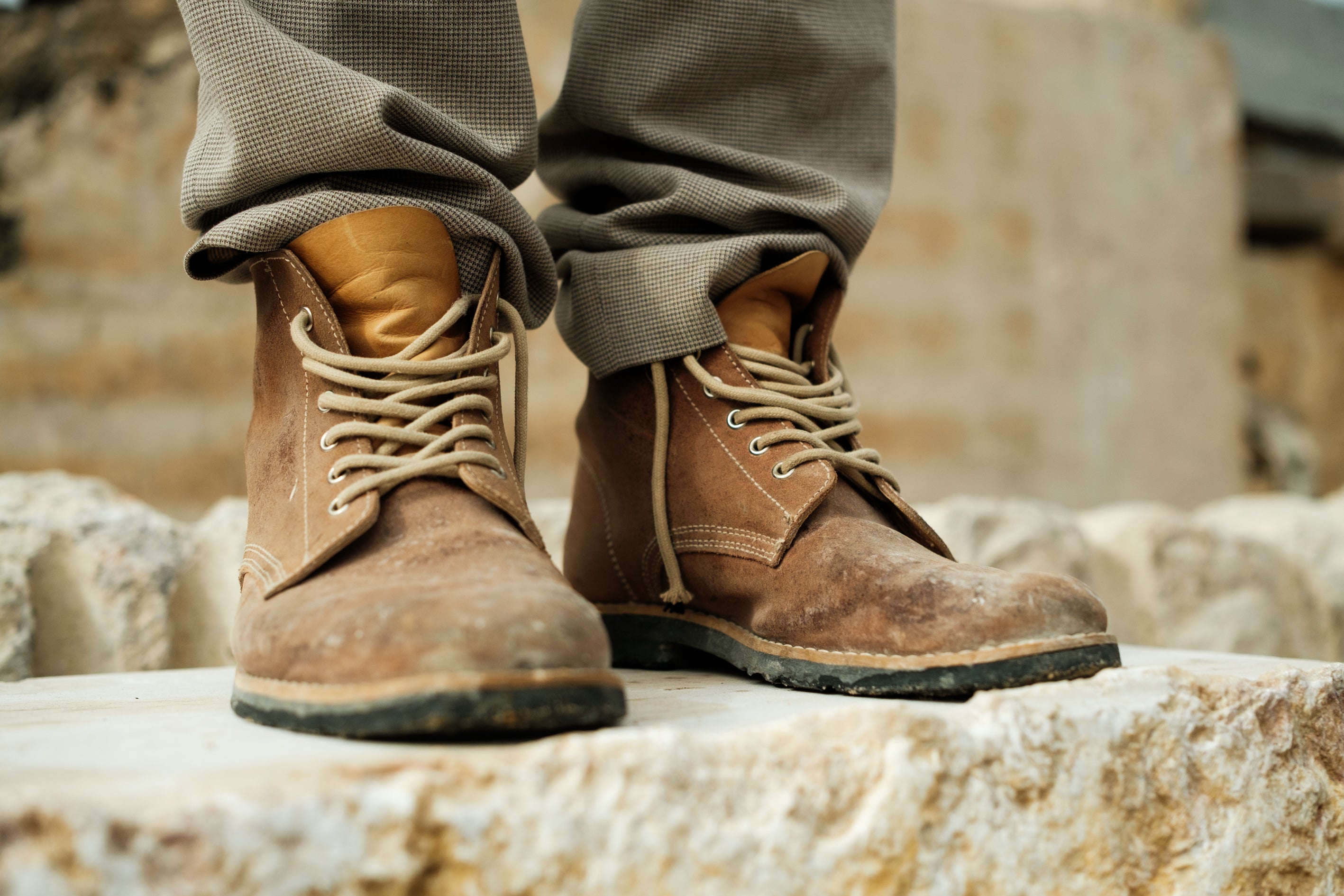 Close-up of Lobo Raw Boots in brown leather, showing natural wear and tear, worn by Carlos Flores standing on a rough stone block. The boots feature beige laces and a rugged patina, highlighting their durability and comfort for architecture and worksite environments.