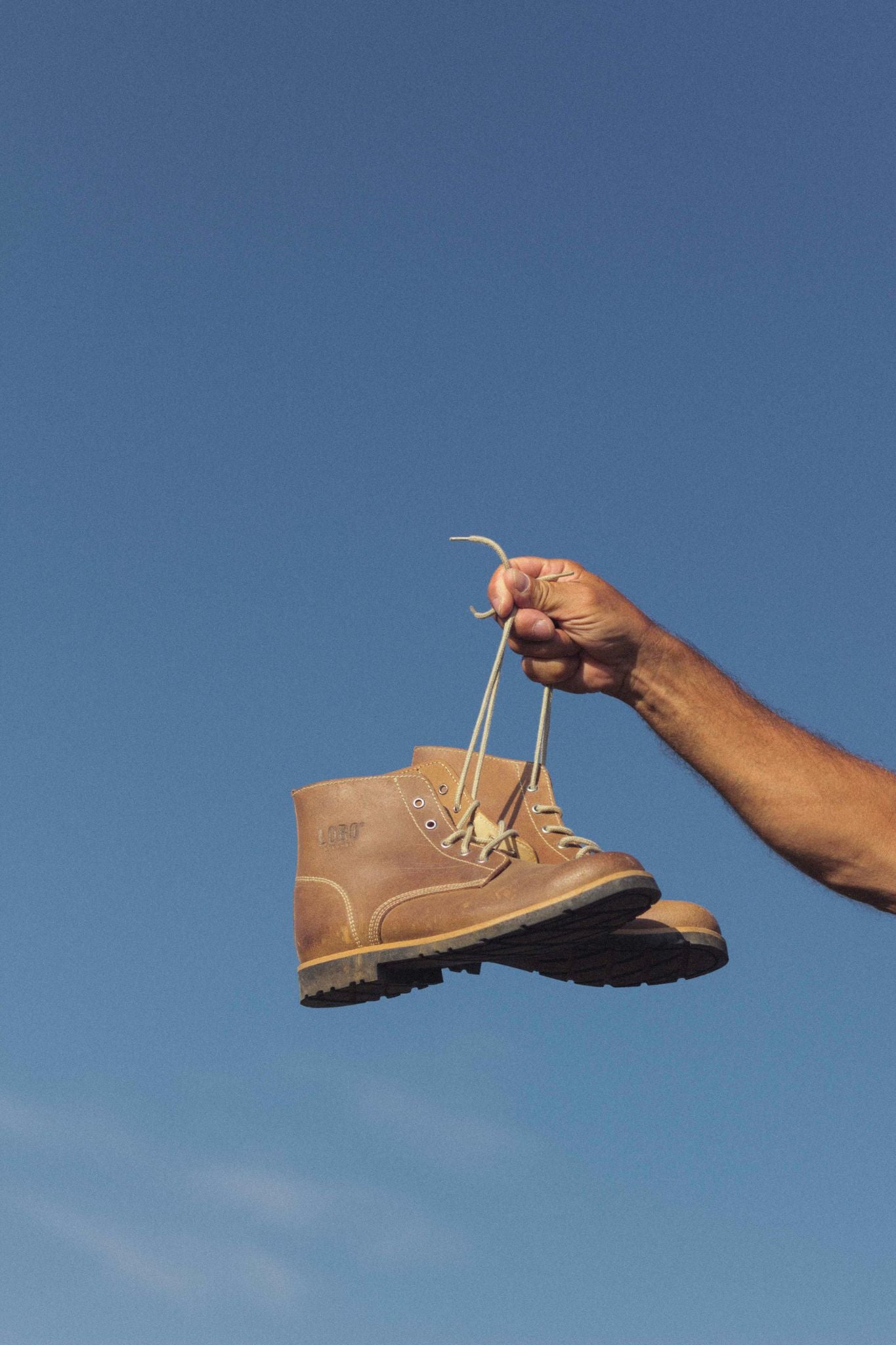 A man's hand holding a pair of natural leather Lobo Raw Boots, highlighting the outdoor and handcrafted style against a blue sky.