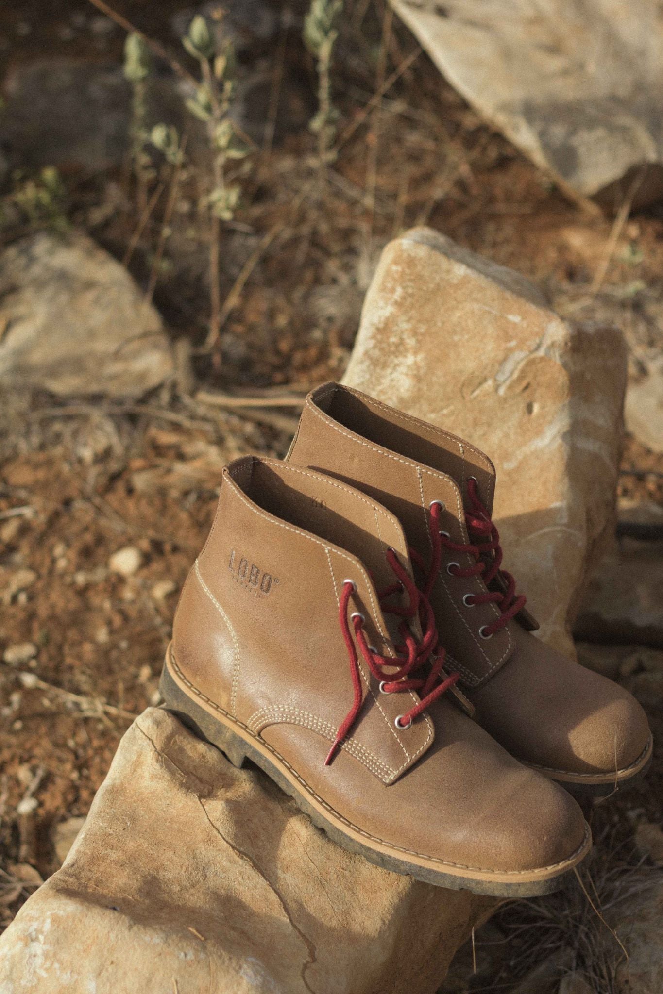 Lobo Raw Boots in brown leather with red laces, placed on rocks in a natural outdoor setting.