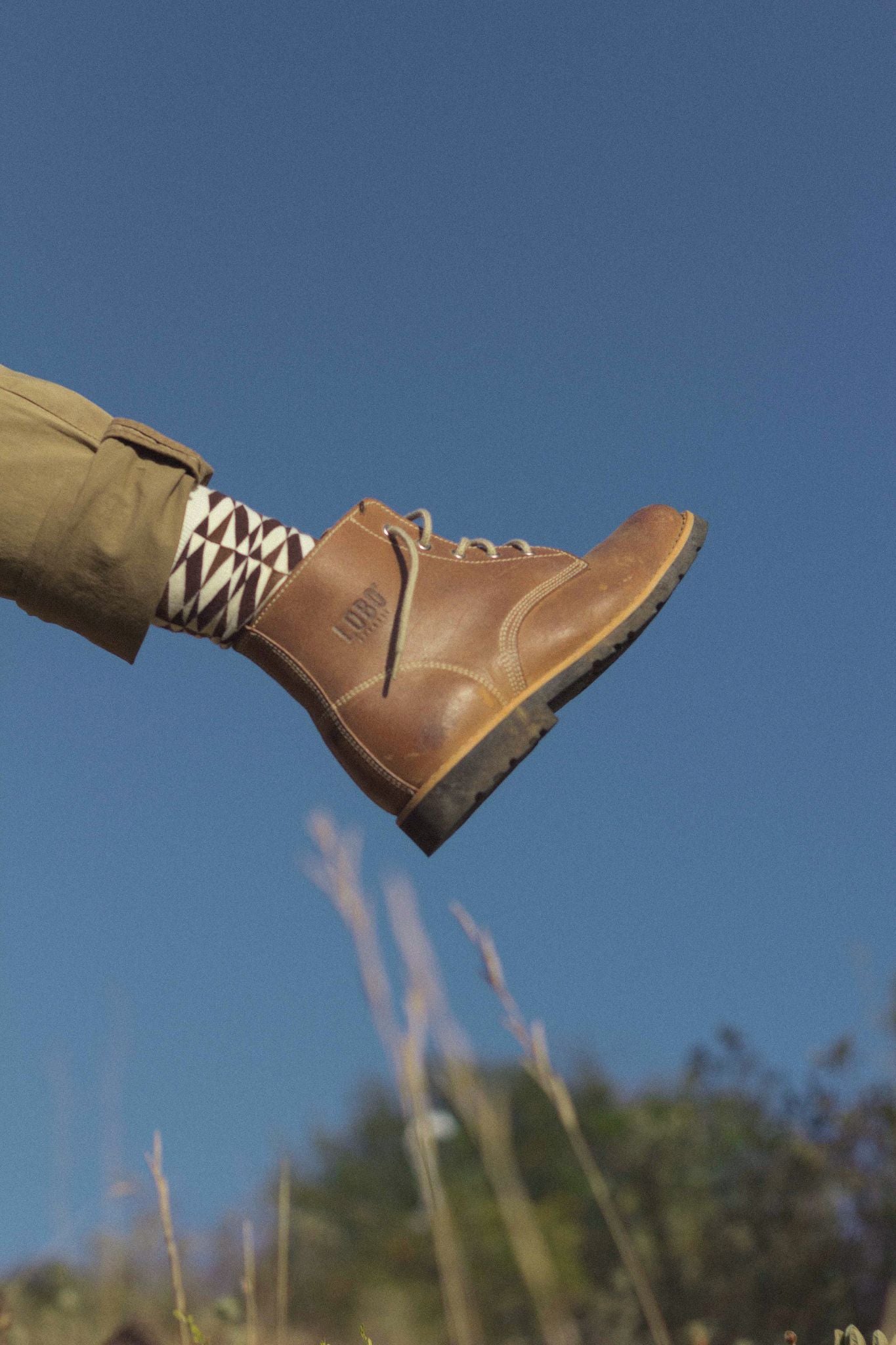 Side view of a Lobo Raw Boot held up against a clear blue sky, styled with geometric patterned socks and khaki chinos.