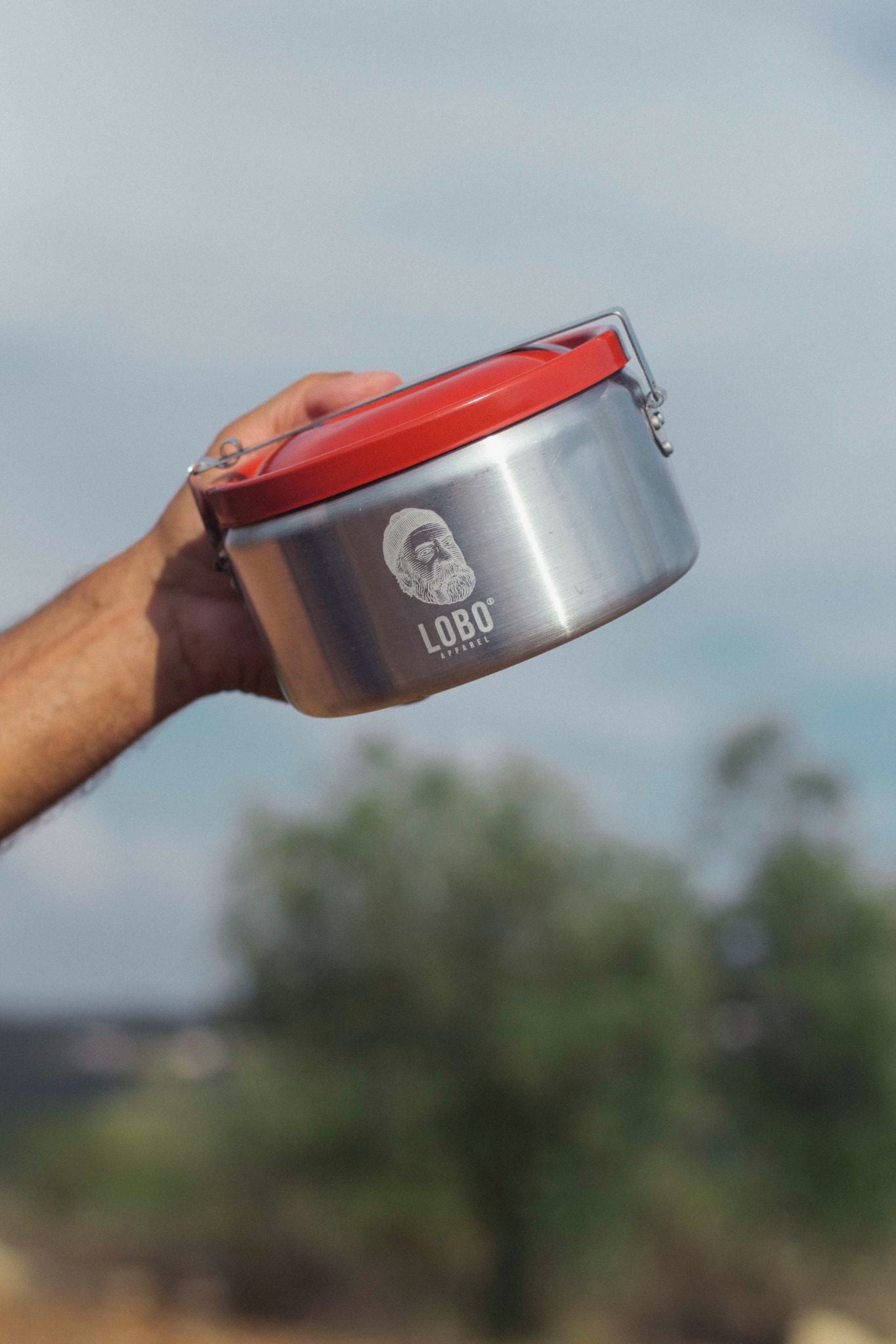 Hand holding an aluminum camping mess tin with a red lid, featuring the "Lobo Apparel" logo and a bearded man illustration, against a blurred nature background.