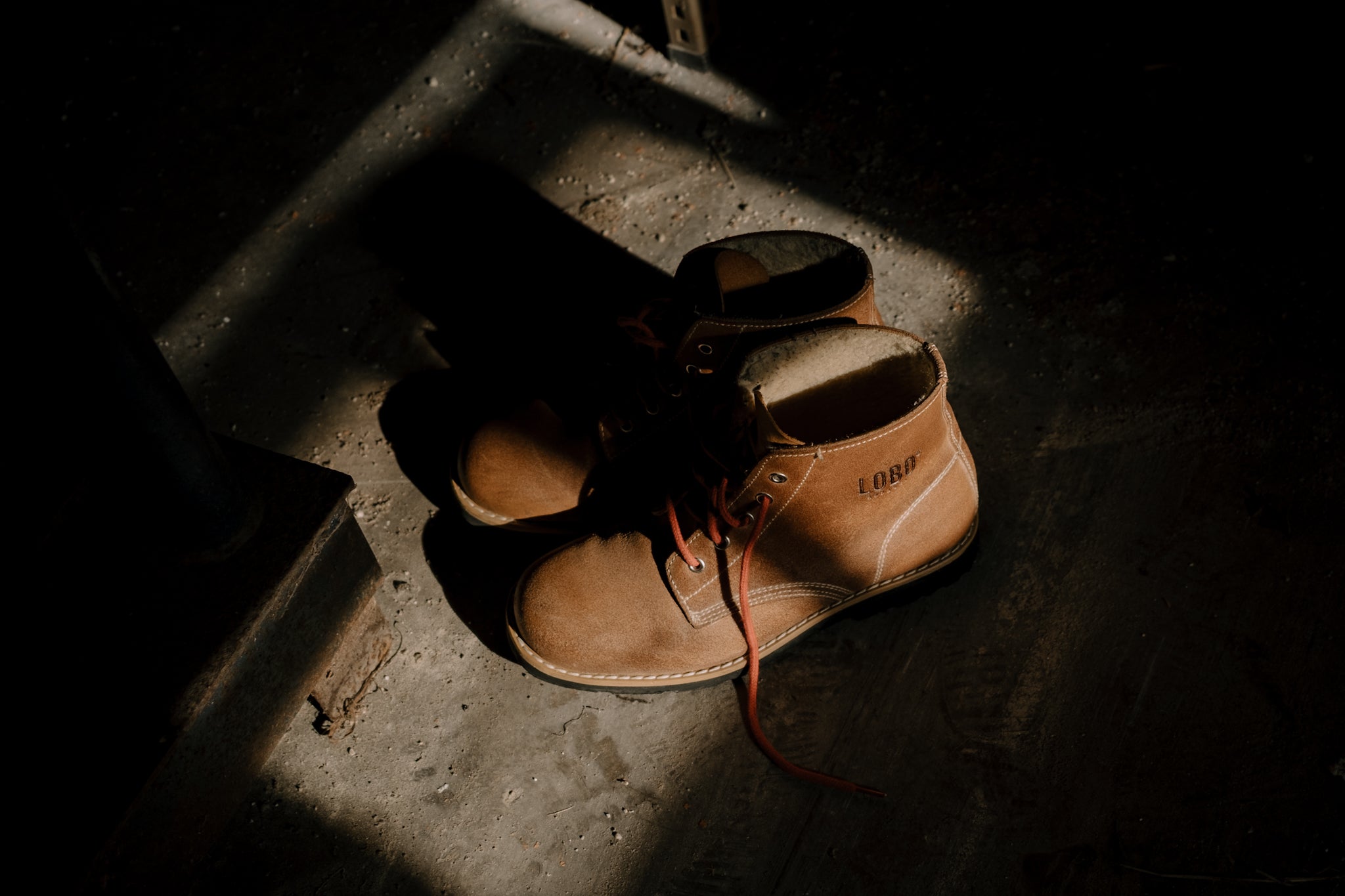 Dramatic high-angle shot of Lobo Raw Boots in leather, placed on a textured concrete floor and illuminated by a lateral beam of sunlight in an old house.