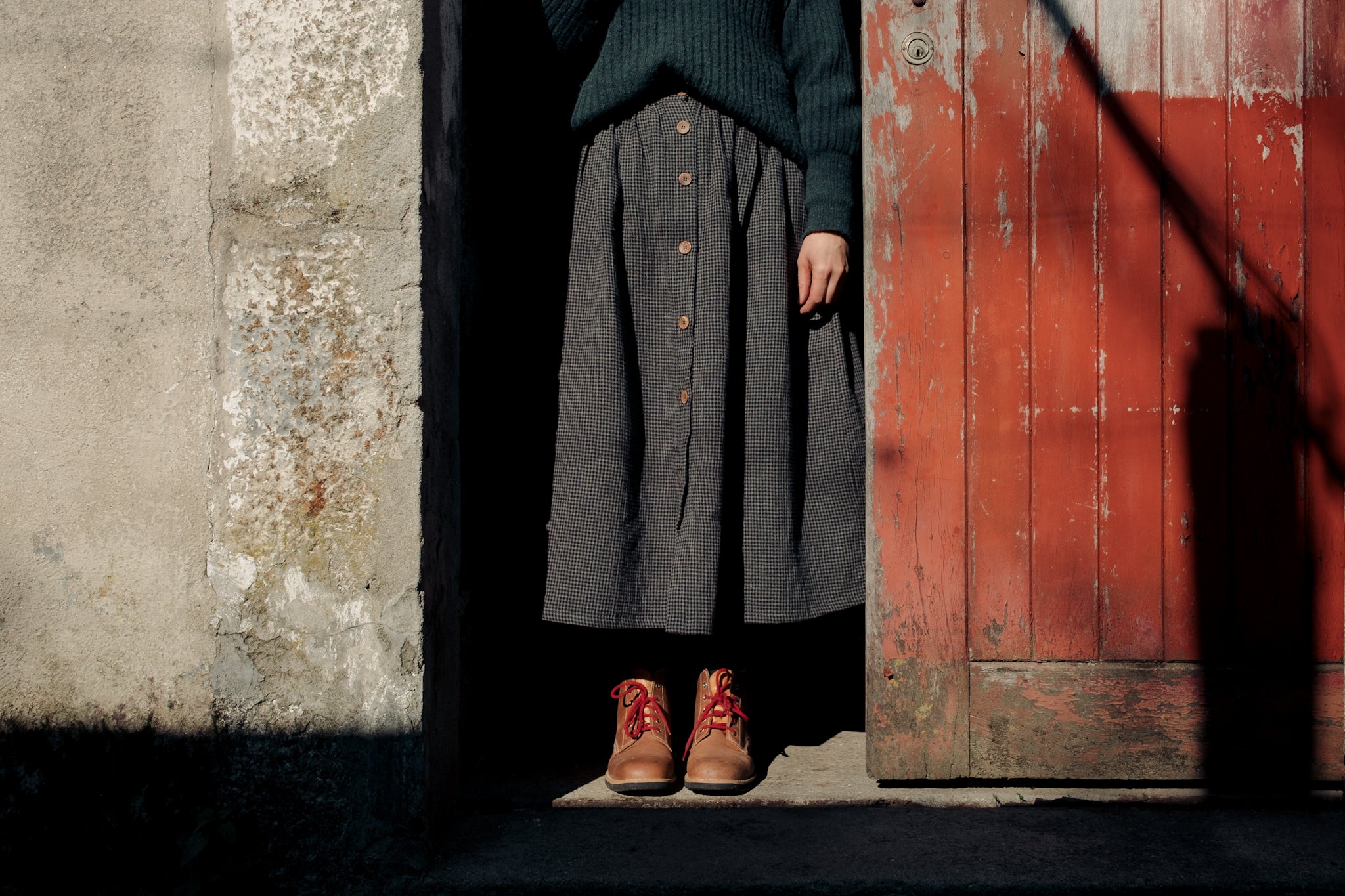 Detail view of an editorial look featuring a midi skirt and knit jumper, paired with Lobo Raw Boots and red laces, framed by a rustic wooden door.