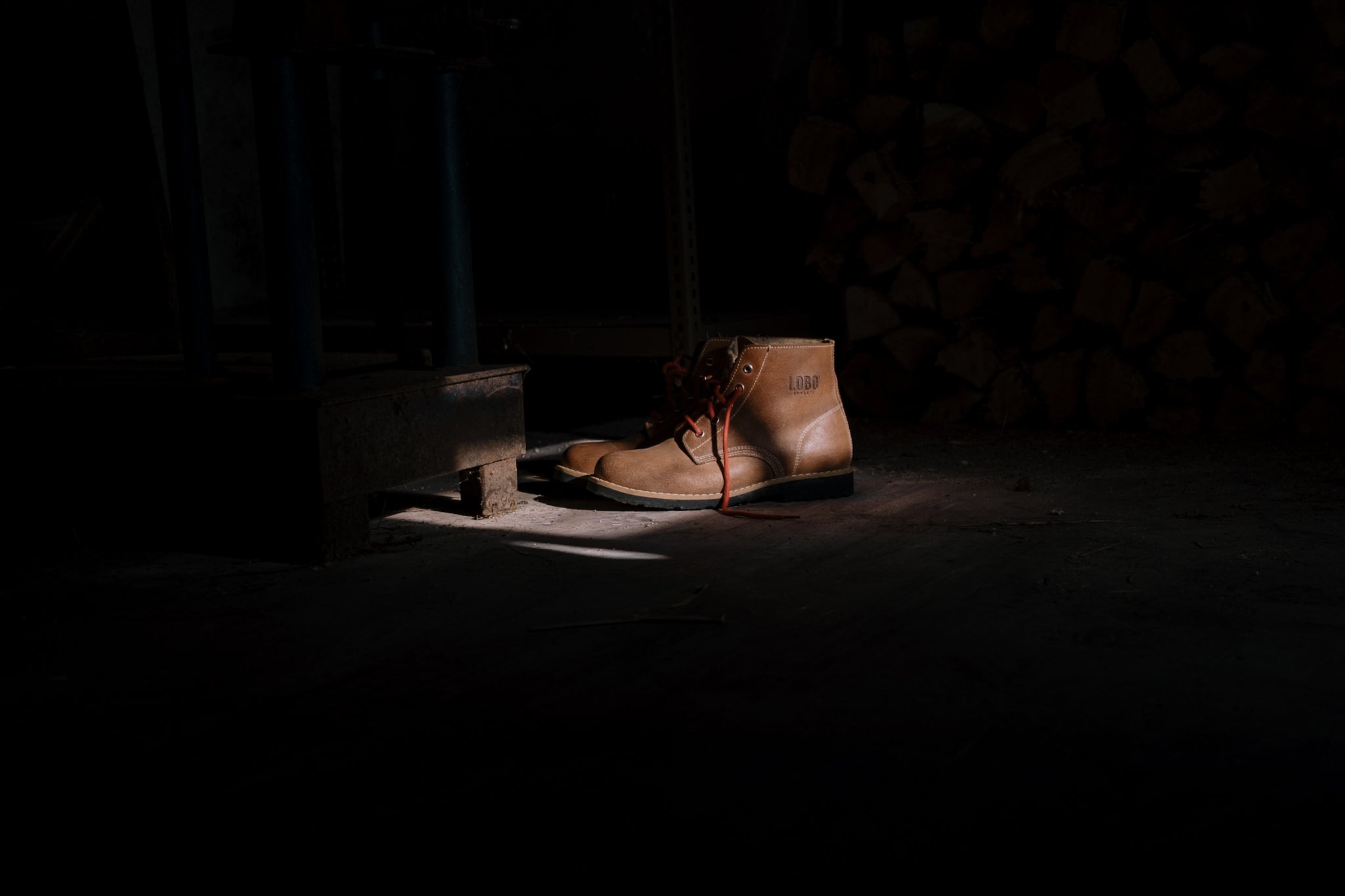 Pair of brown leather Lobo Raw Boots with red laces, highlighted by natural light in a rustic and moody cottage setting.
