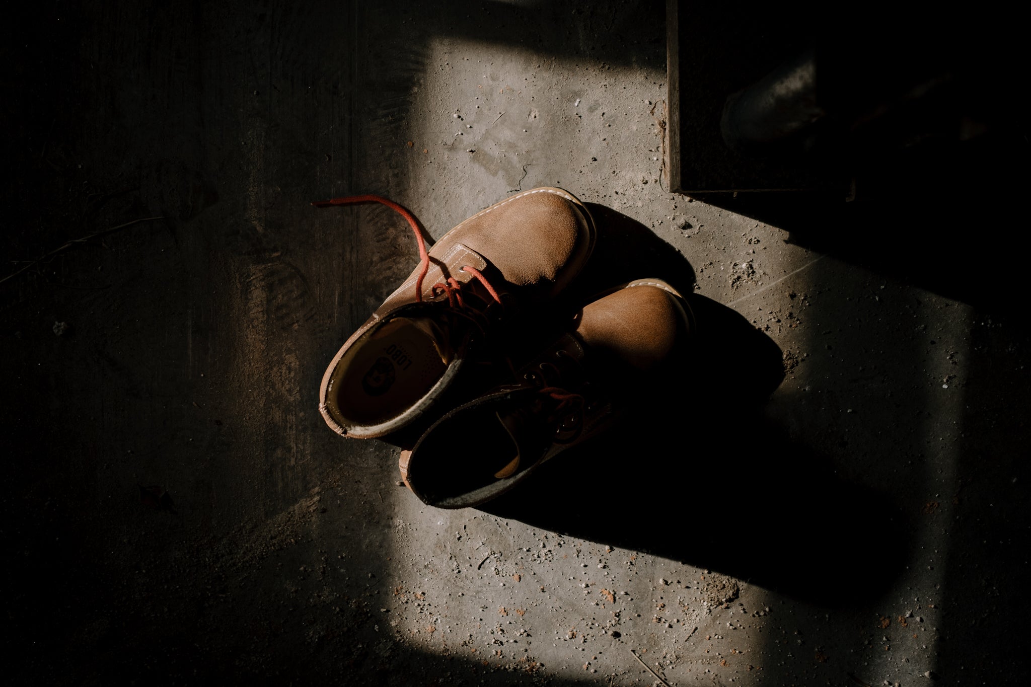 Flat lay top view of Lobo Raw Boots in brown leather, resting on textured concrete in a moody setting with direct sunlight.