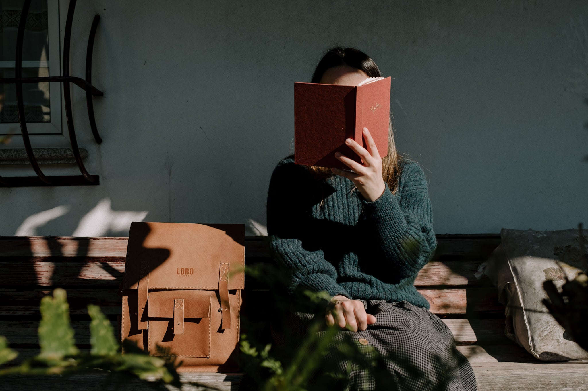 Front view of a woman seated on a rustic wooden bench, reading a red-cover book. She wears a dark green knit sweater and a textured skirt. Beside her is a handcrafted Lobo leather backpack. The scene is bathed in natural light and direct shadows, conveying serenity and the Portuguese slow-living concept.