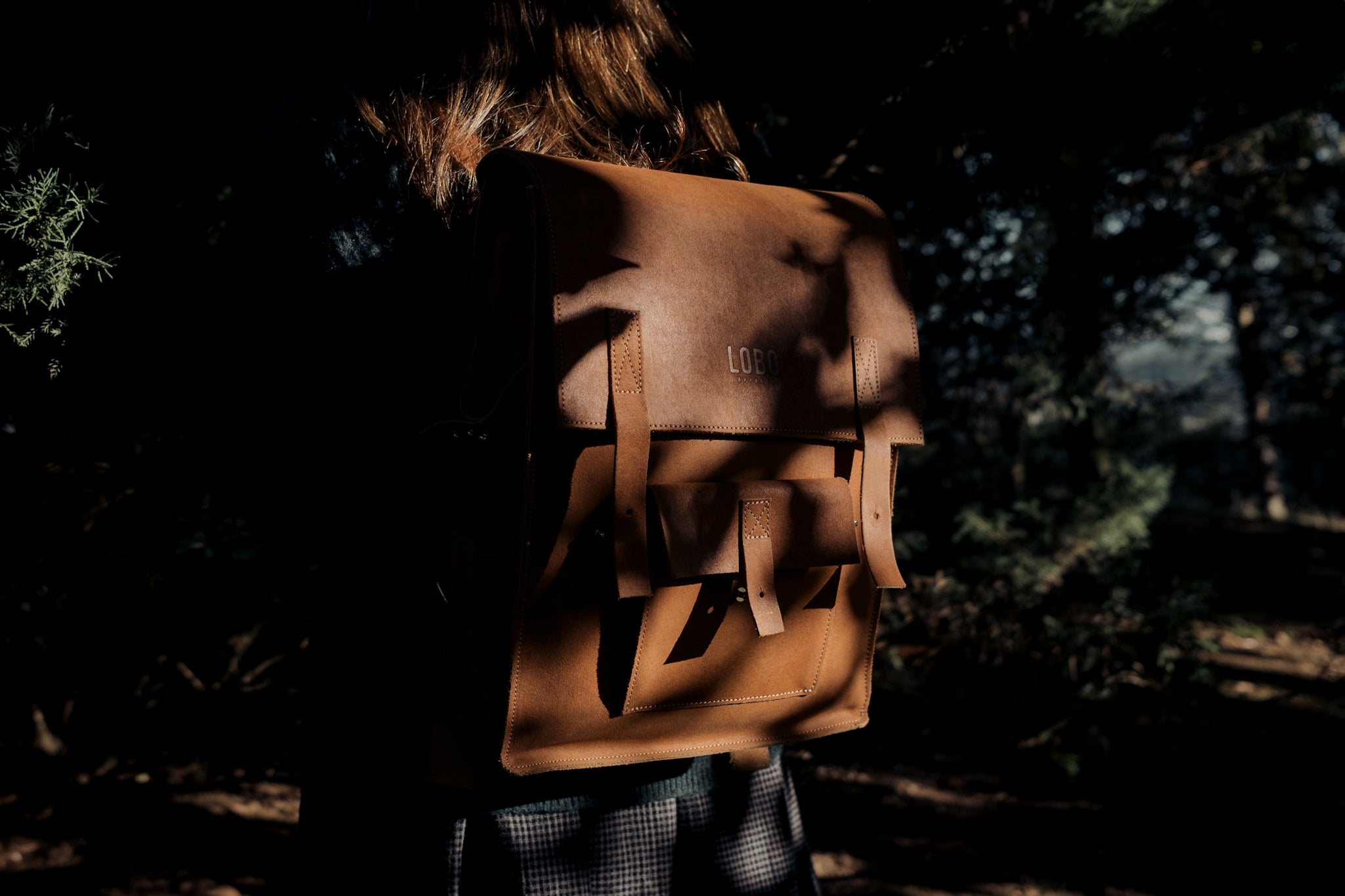 Back medium shot of a woman wearing a natural-colored Lobo leather backpack, framed by the deep shadows and light of a forest.
