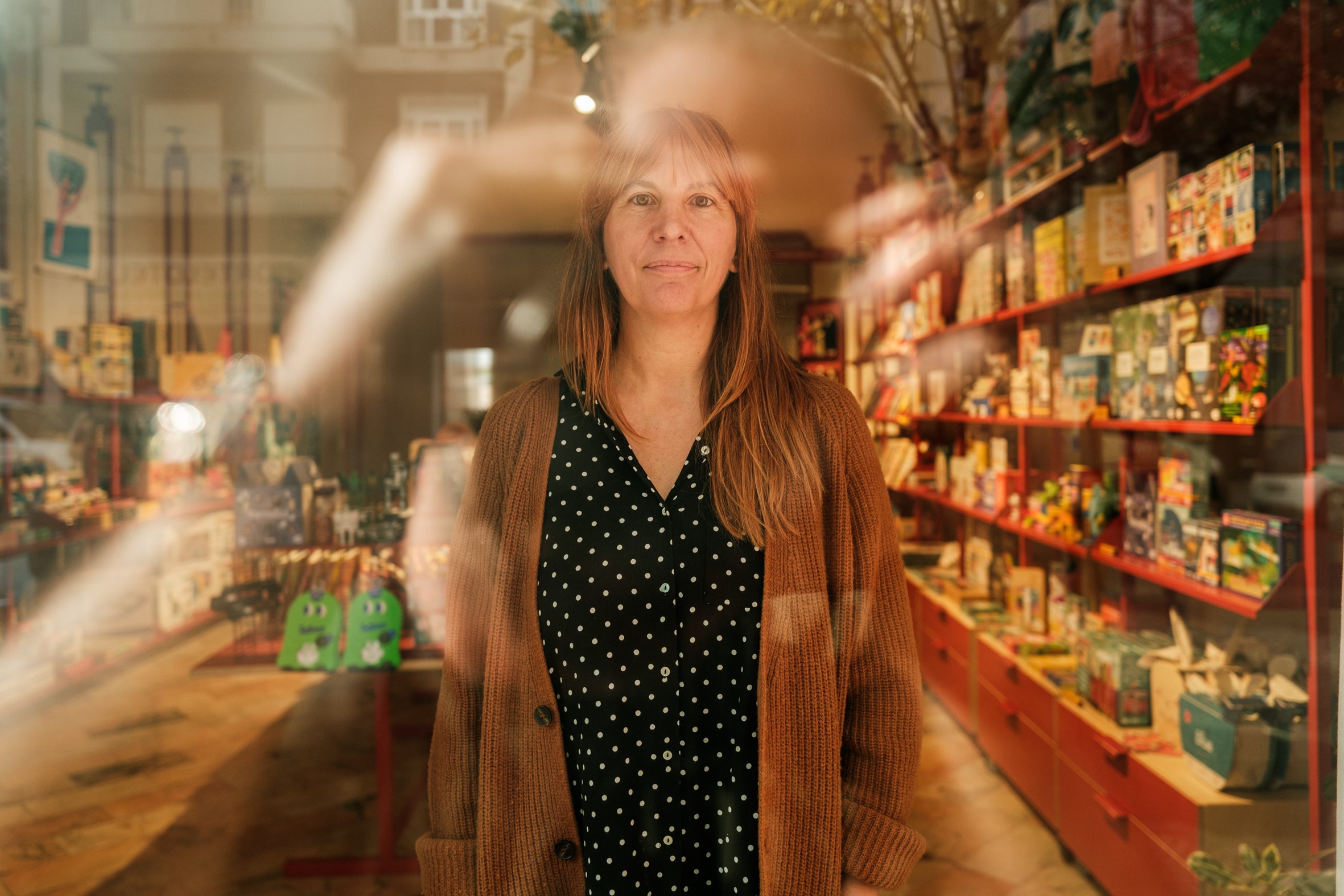Portrait of the creator of Fulana, Beltrana e Cicrana, seen through a window reflection inside her Fufu shop and studio. She is wearing the Lobo Brown Wool Cardigan over a polka-dot shirt, surrounded by shelves filled with toys, books, and design objects. The image highlights the versatility and comfort of Lobo knitwear in an artist's daily life.