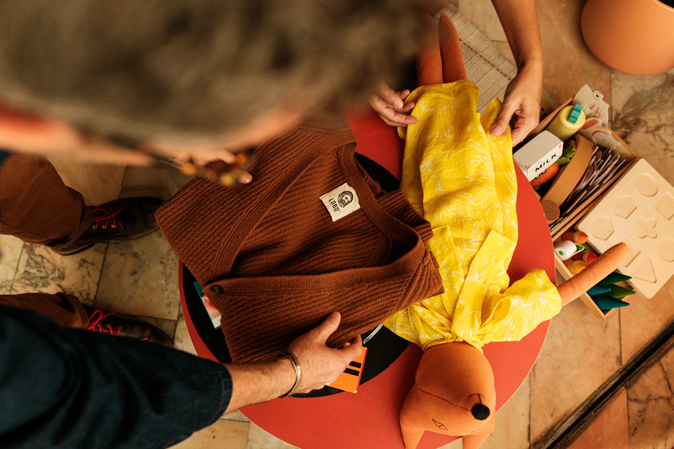 Top view of a Lobo Brown Wool Cardigan folded on a red round table, next to a handmade cloth doll by "Fulana, Beltrana e Cicrana". The white LOBO label is visible, featuring a bearded man wearing a beanie and smoking a pipe. The scene highlights comfort, premium wool quality, and the intersection of fashion design and contemporary crafts.