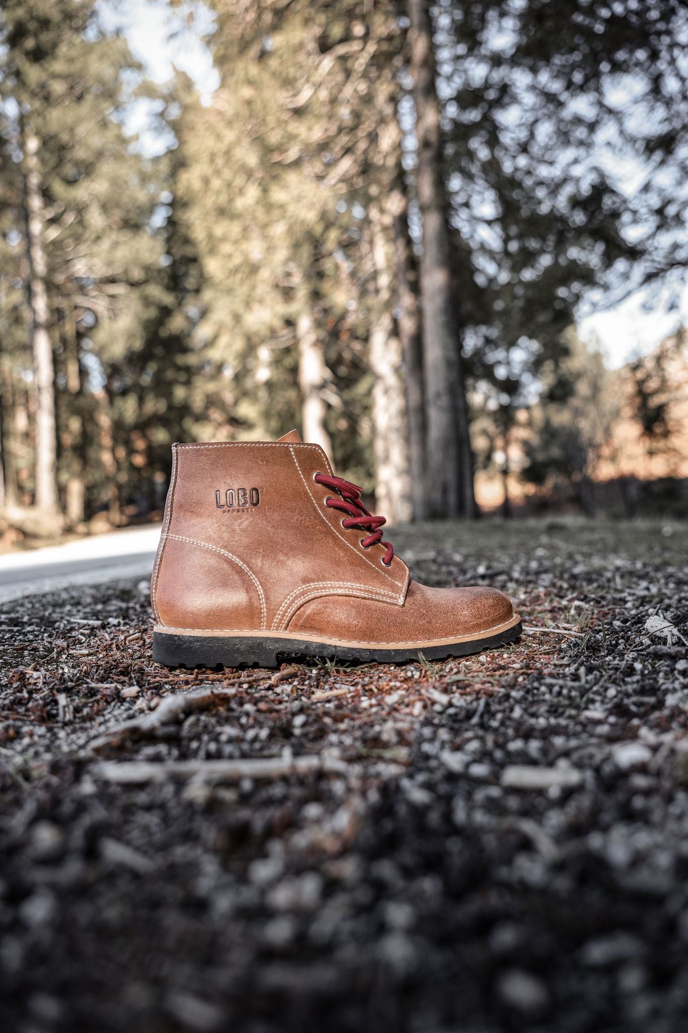 Side view of brown leather Lobo Raw Boots with red laces, placed on a forest floor of pine needles and dirt, with a blurred pine forest in the background.