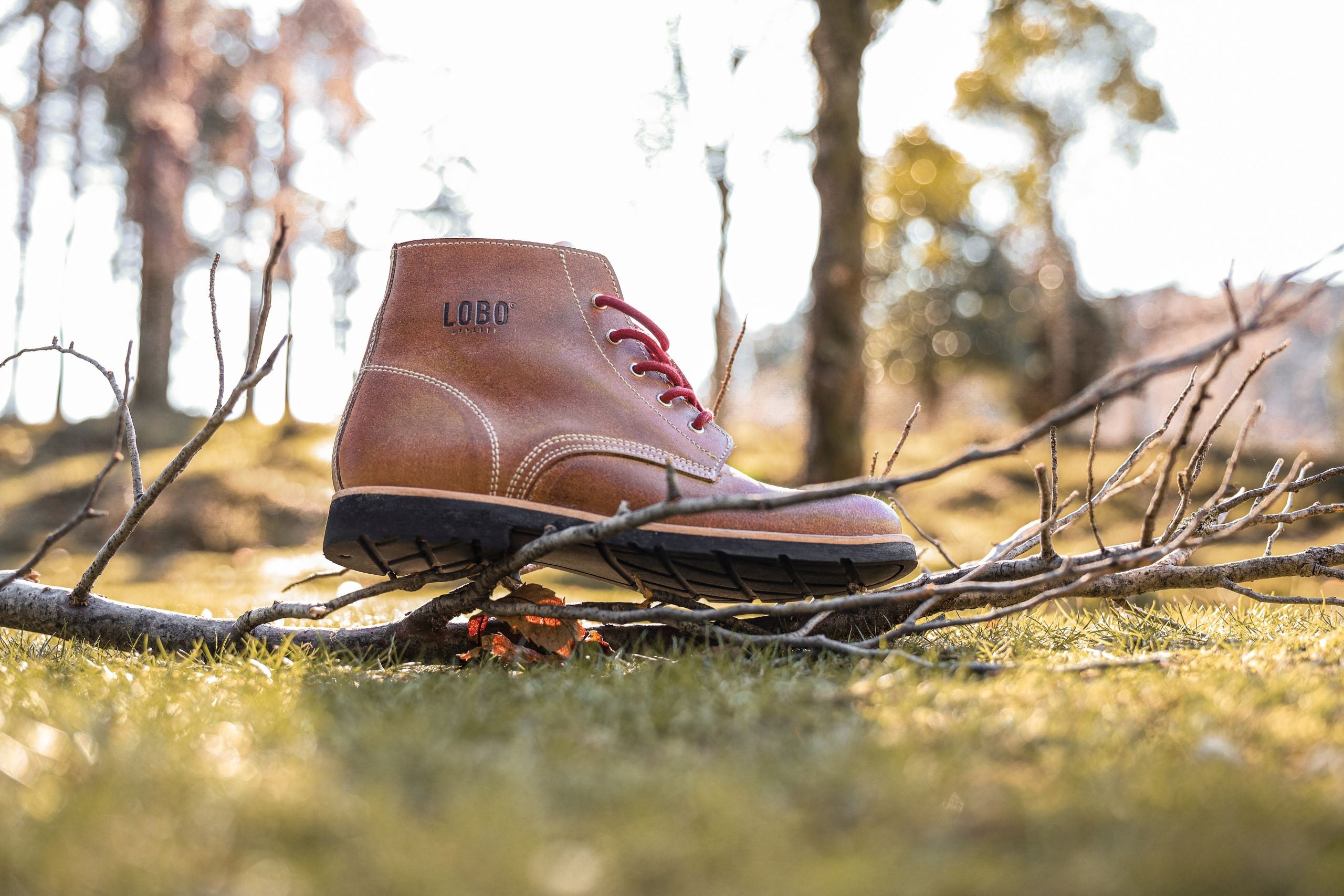 Lobo Raw Boots in brown leather balanced on dry branches, showcasing the rugged black sole and the embossed logo on the side.