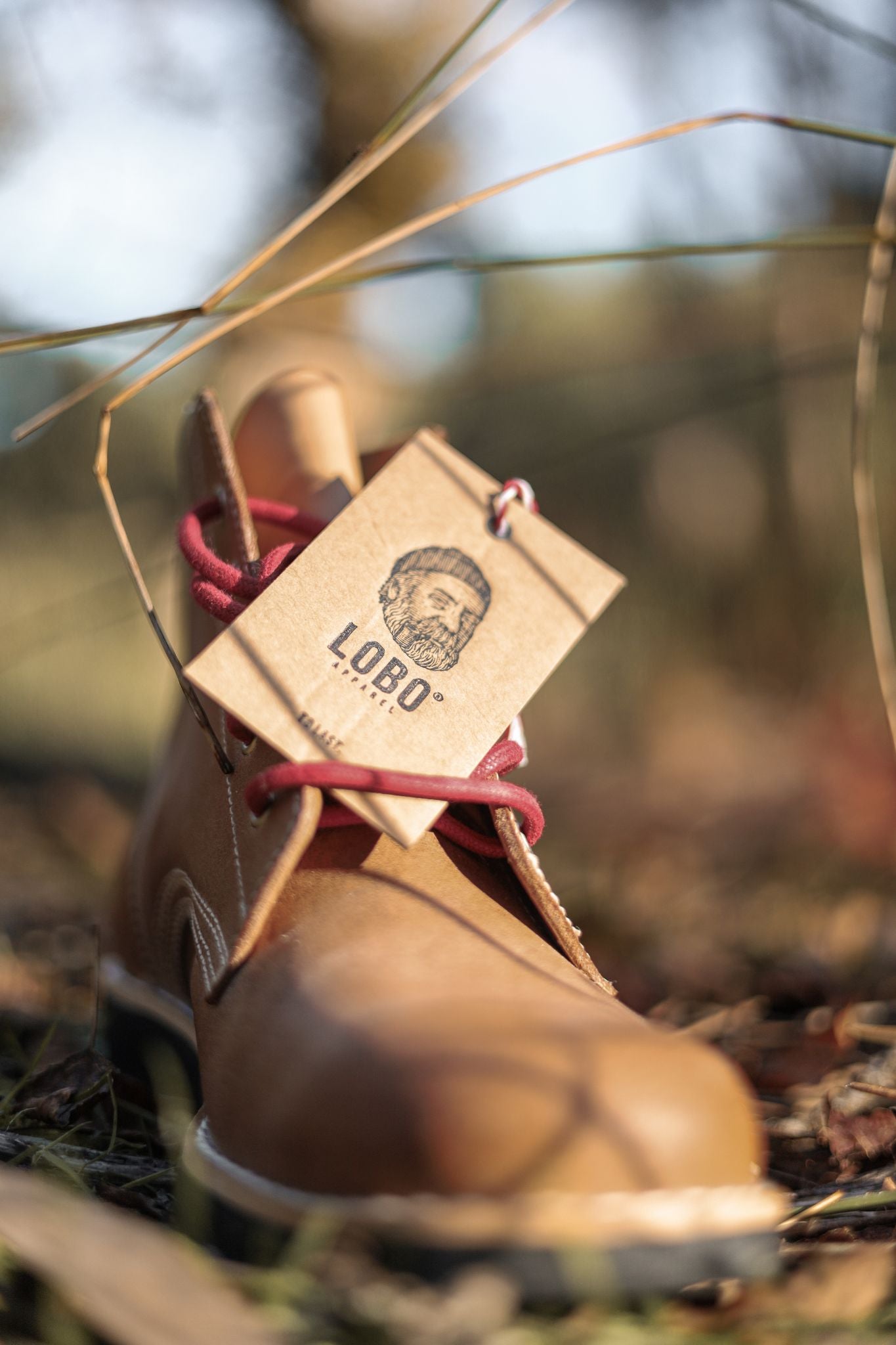 Close-up of the Lobo Apparel cardboard hangtag attached to a brown leather boot with red laces. The tag displays the official logo, highlighting the handcrafted footwear concept.