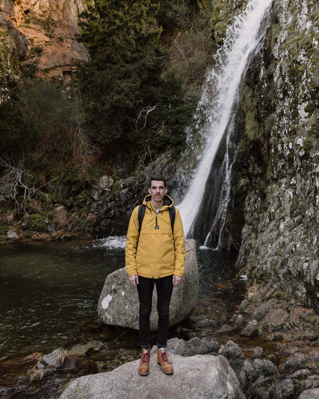Man wearing a technical yellow jacket and backpack, standing in front of a natural waterfall, representing the spirit of adventure and the practical use of the Lobo Apparel collection