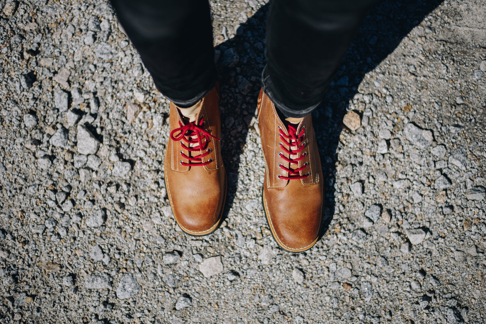 Top-down view of the Lobo Raw Boots being worn by a person in black trousers, standing on a gravel path, highlighting the pattern of the red laces and the boot's rounded toe shape.