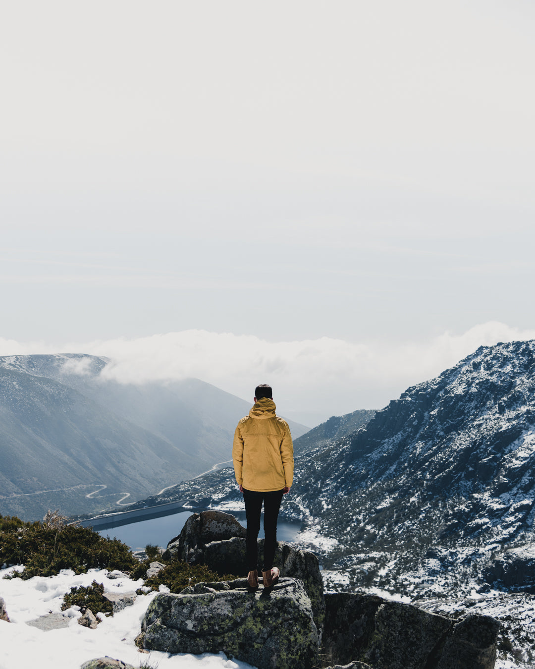 Explorer wearing a Lobo Apparel yellow parka on a frozen mountain peak, overlooking a dam valley, symbolizing achievement and the brand's outdoor spirit.