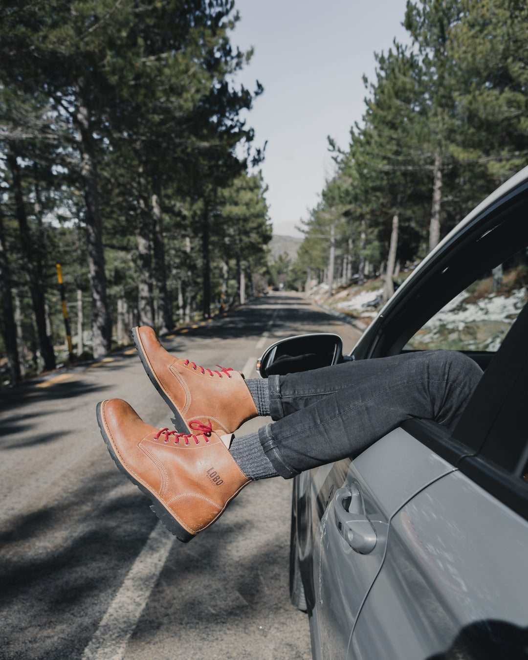 Lobo Raw Boots showcased with legs out of a car window on a mountain road lined with pine trees, evoking freedom and adventure.