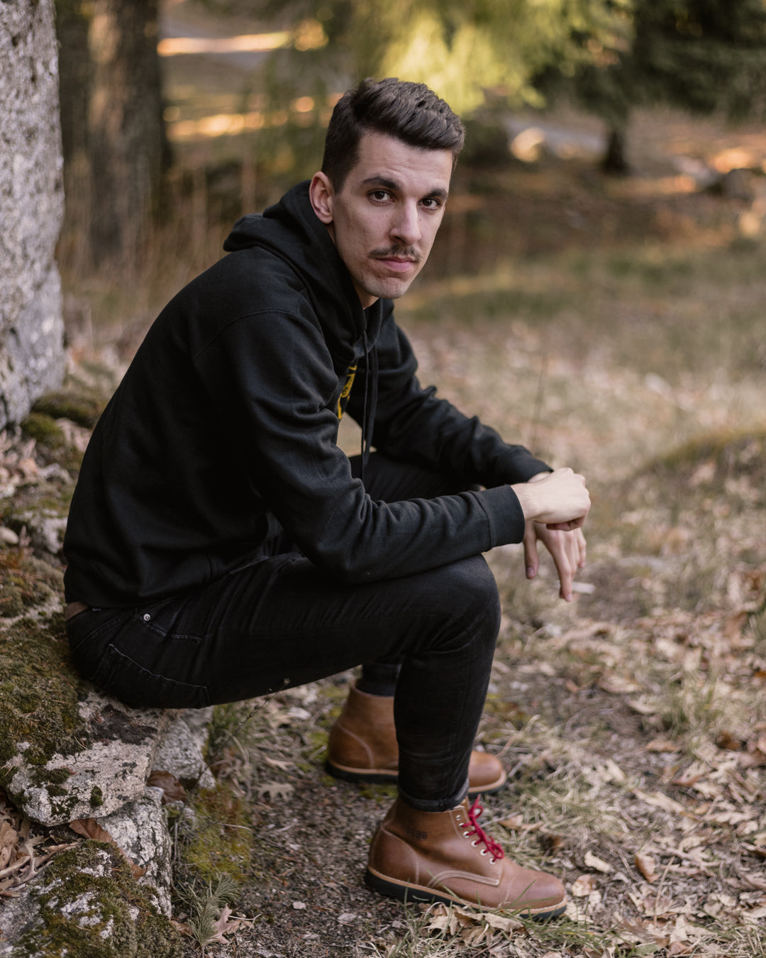 Portrait of a man wearing a black hoodie and dark trousers, featuring Lobo Raw leather boots, sitting in an autumnal forest setting.