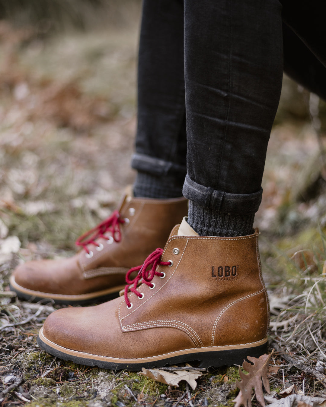 Close-up side view of the Lobo Raw Boot in brown leather, showcasing the contrast white stitching, the embossed "LOBO" logo, and vibrant red laces in a natural outdoor setting.