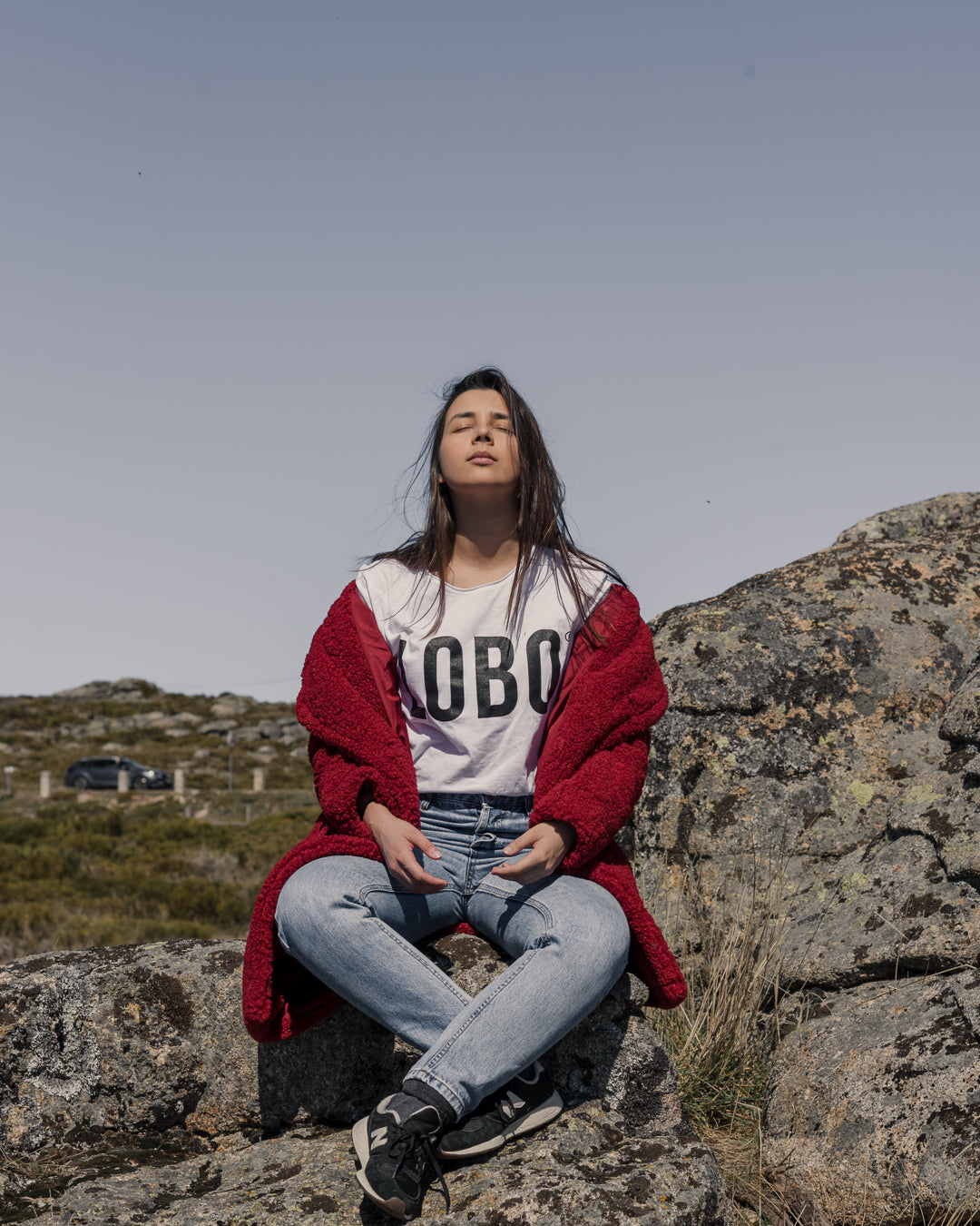 Mulher sentada numa rocha de montanha com t-shirt Lobo branca de logótipo grande, casaco bordeaux e calças de ganga, transmitindo serenidade e conforto outdoor.