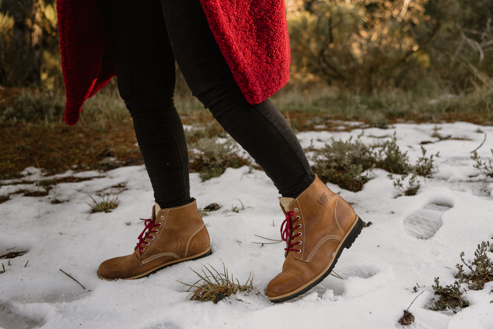 Close-up of Lobo Raw brown leather boots walking on snow, paired with black trousers and a red coat, highlighting insulation and style in cold weather.