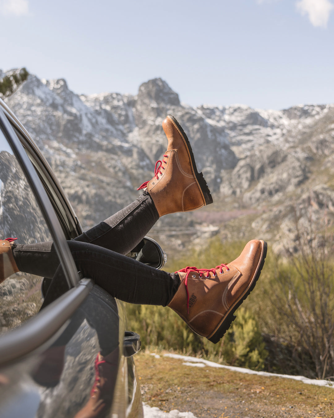 Person relaxing in a car wearing Lobo Raw Boots, overlooking frozen mountains, evoking a spirit of adventure and travel.