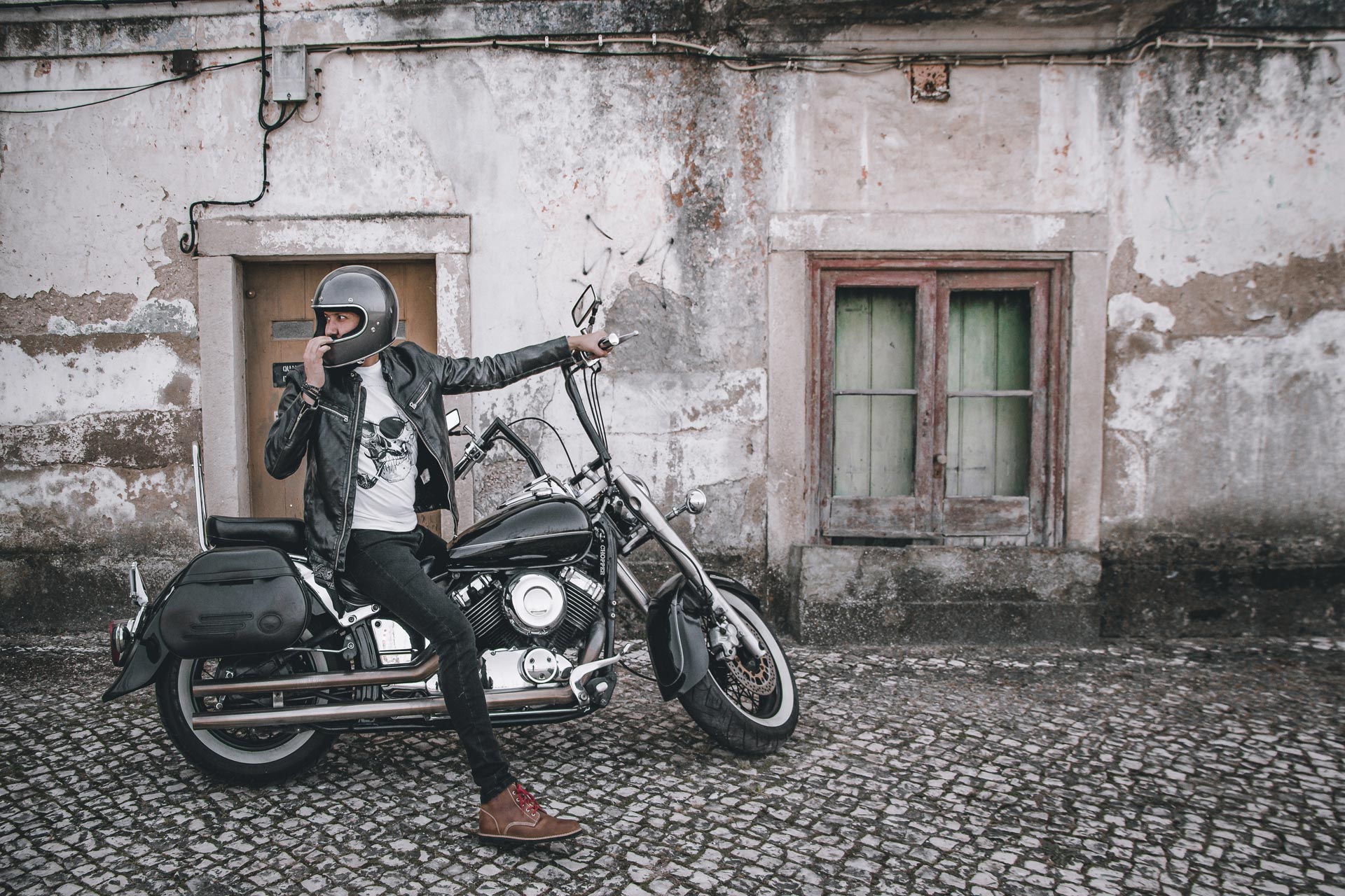 Motorcyclist in a helmet and leather jacket, seated on a classic custom motorcycle with ape-hanger handlebars, wearing Lobo Raw boots with red laces, in an old urban setting with Portuguese cobblestones and a rustic facade.