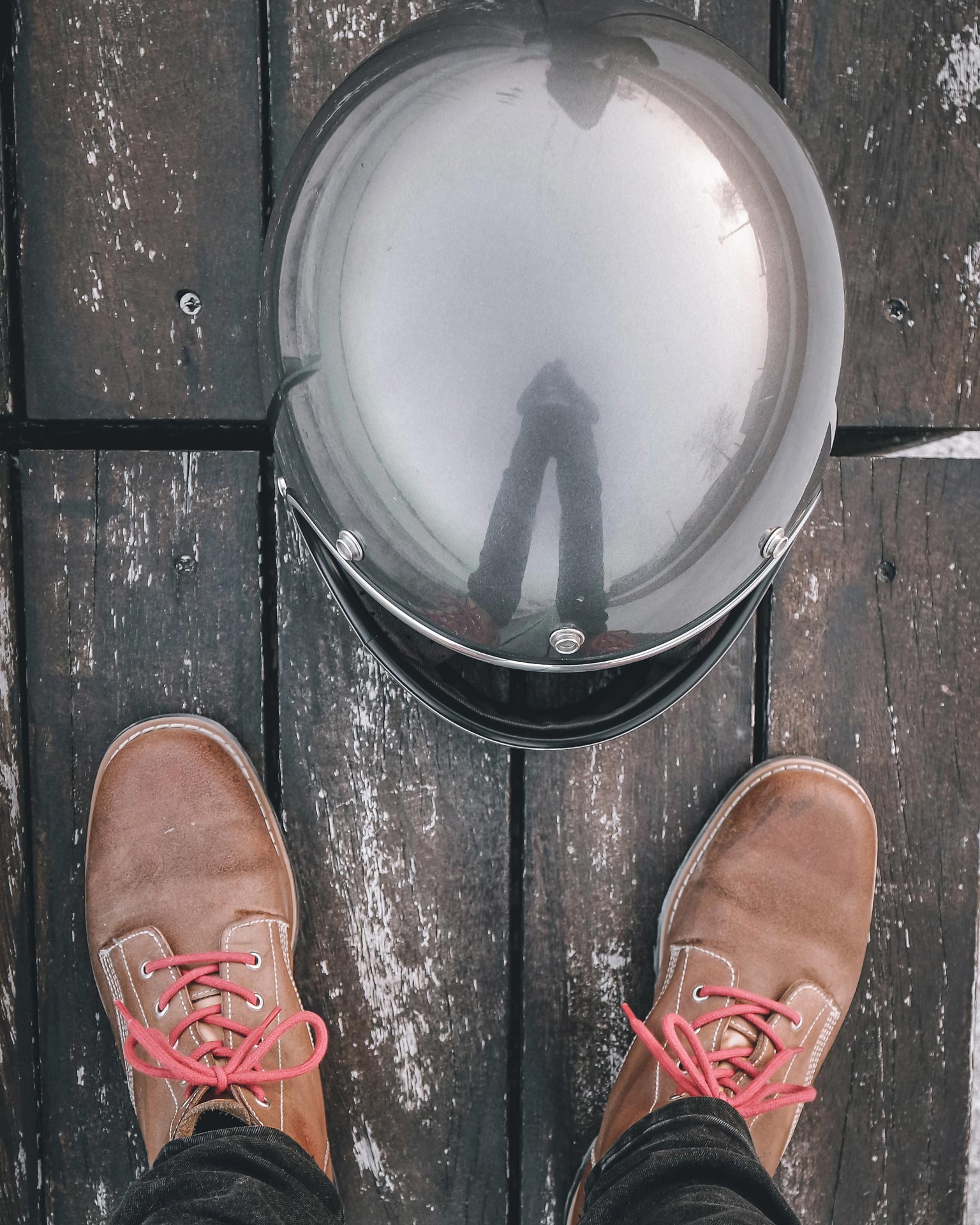 Top-down flat-lay view of Lobo Raw leather boots with red laces, positioned next to a matte black motorcycle helmet, all on weathered wooden planks.