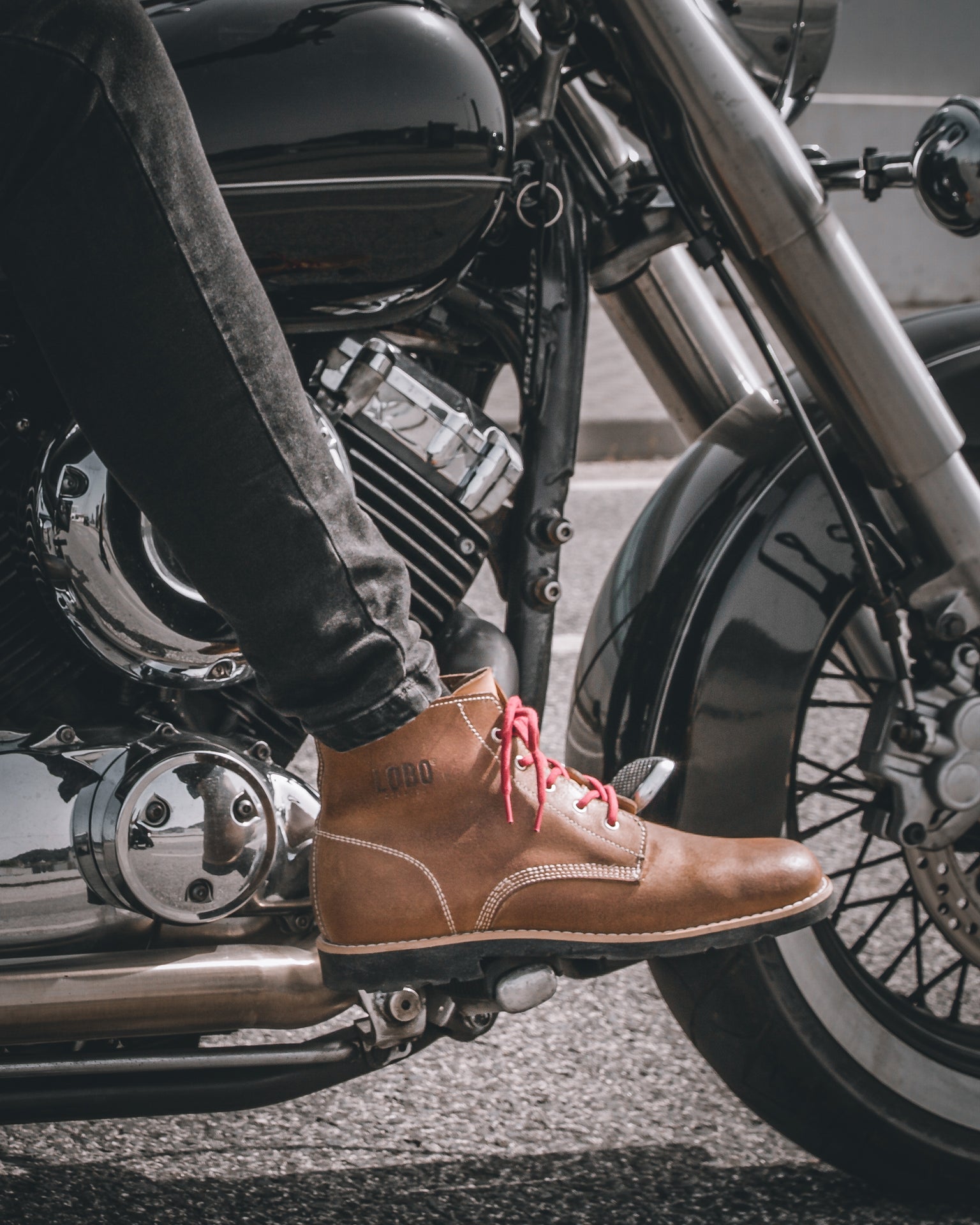 Lobo Raw brown leather boots with vibrant red laces, with the foot positioned on a classic custom motorcycle's footrest with a chrome engine, showcasing road durability.