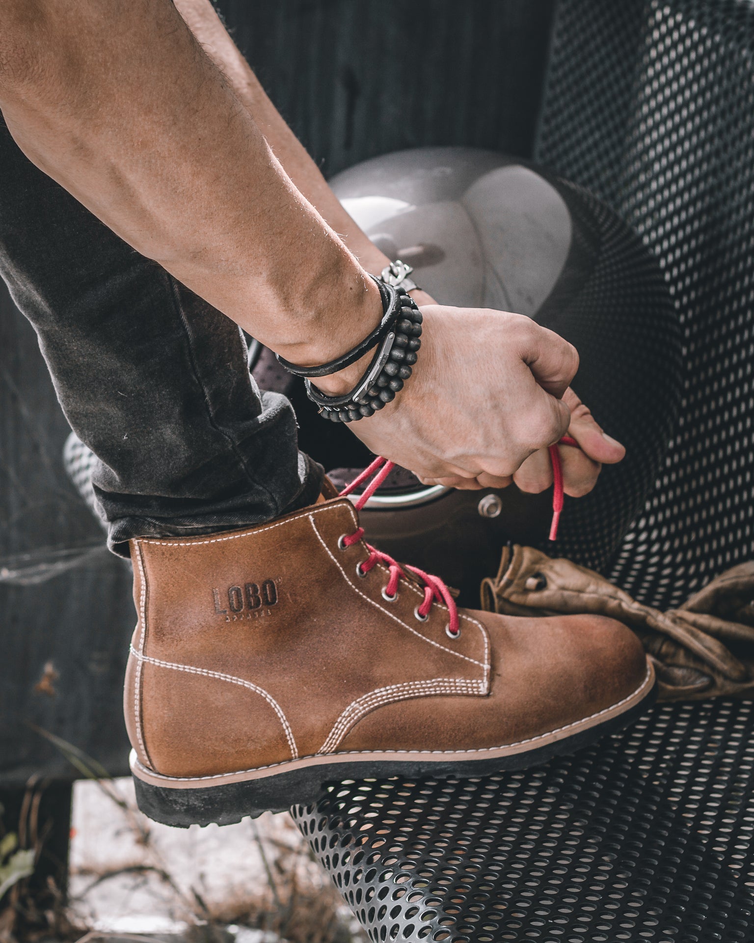 Close-up of hands tying the red laces of a Lobo Raw boot, with a matte black motorcycle helmet and leather gloves in the background, conveying the moment of journey preparation.