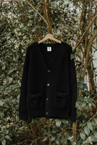 Full view of the black Lobo Black Wool Cardigan on a hanger, with front buttons and patch pockets in a natural setting
