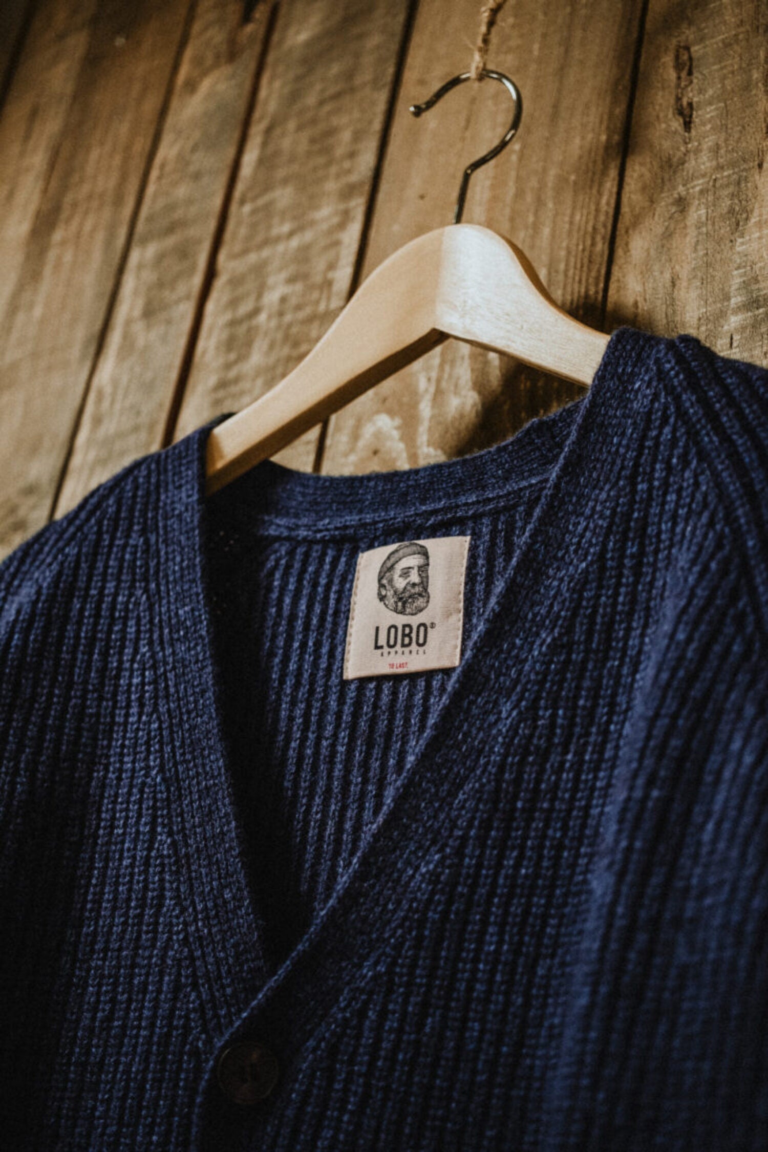 Top detail of the denim blue cardigan on a wooden hanger, showing the Lobo Apparel label and V-neckline.