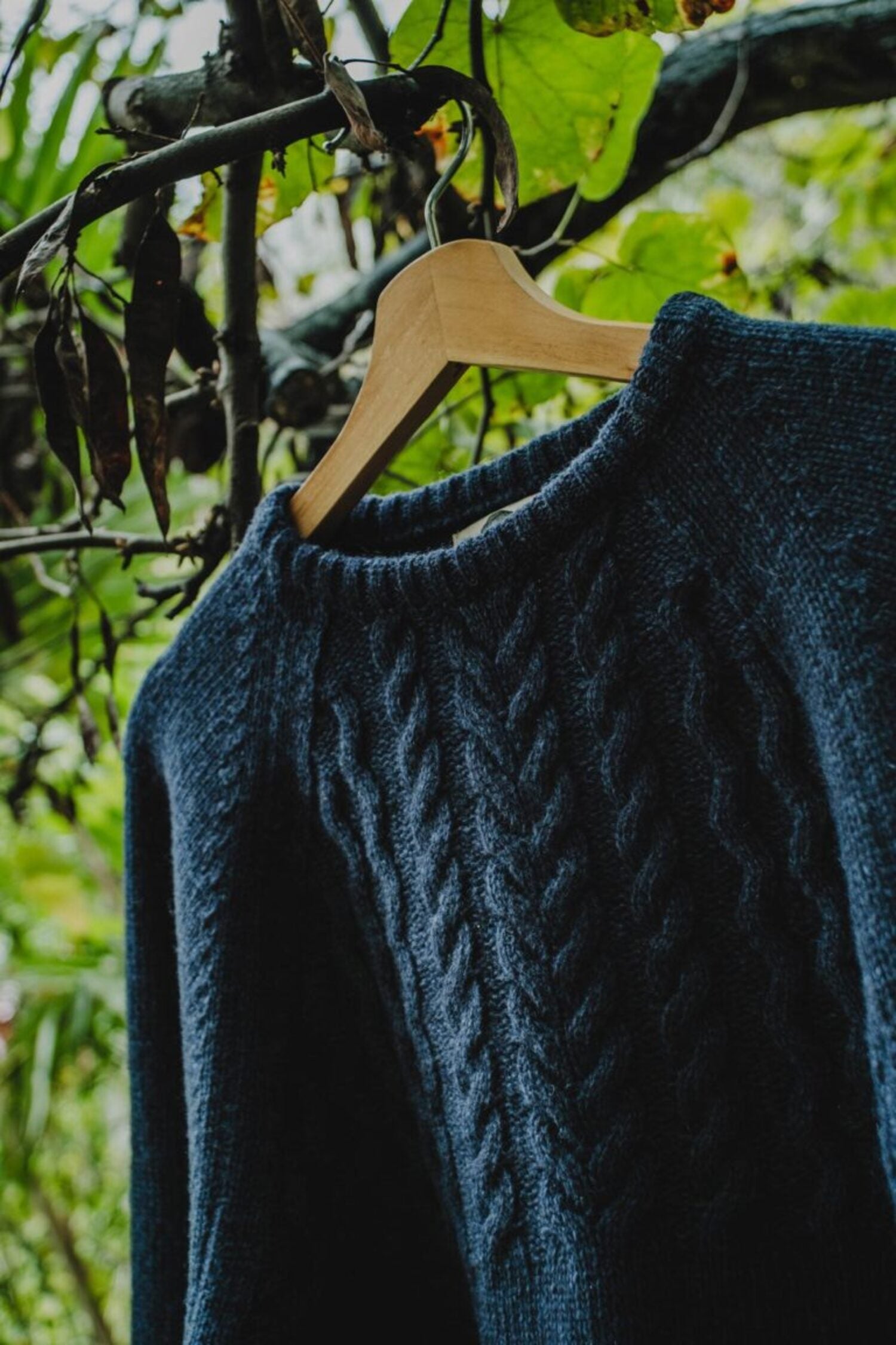 Lobo Blue Fisherman wool sweater in denim blue on a wooden hanger, highlighting the braided knit panel on the chest.