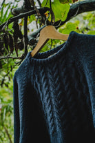 Lobo Blue Fisherman wool sweater in denim blue on a wooden hanger, highlighting the braided knit panel on the chest.