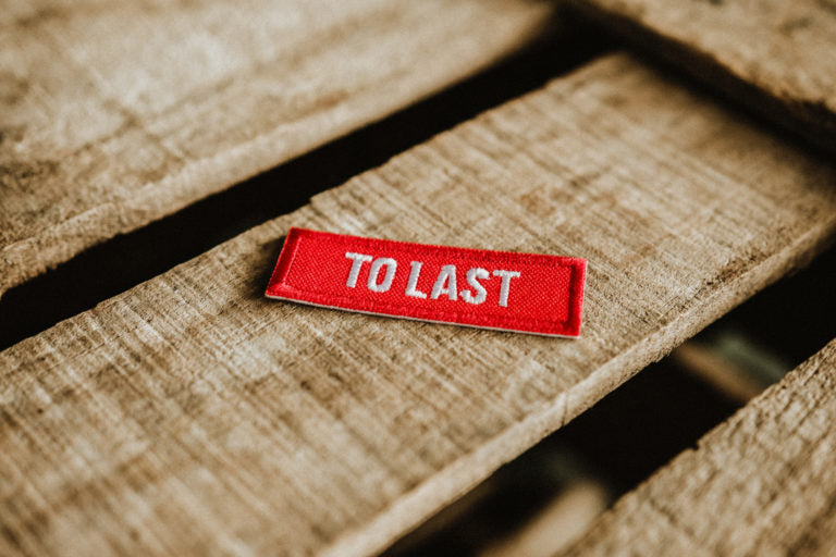 Rectangular embroidered Lobo To Last patch with red background and white "TO LAST" text, resting on a wooden surface.