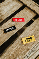 Three Lobo Apparel embroidered patches on a wooden pallet: red "To Last" patch, black "To Resist" patch and yellow Lobo logo patch.