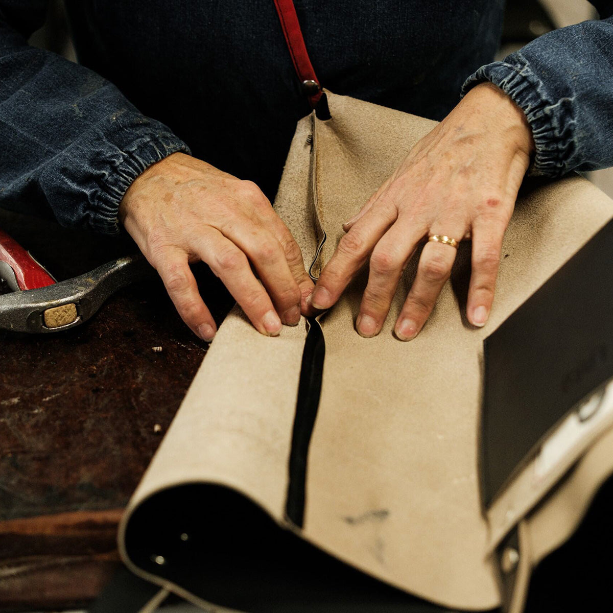 Close-up of a craftsman's hands shaping and working on a beige leather piece on a workbench, with a hammer nearby. Detail of the handmade leather crafting process.