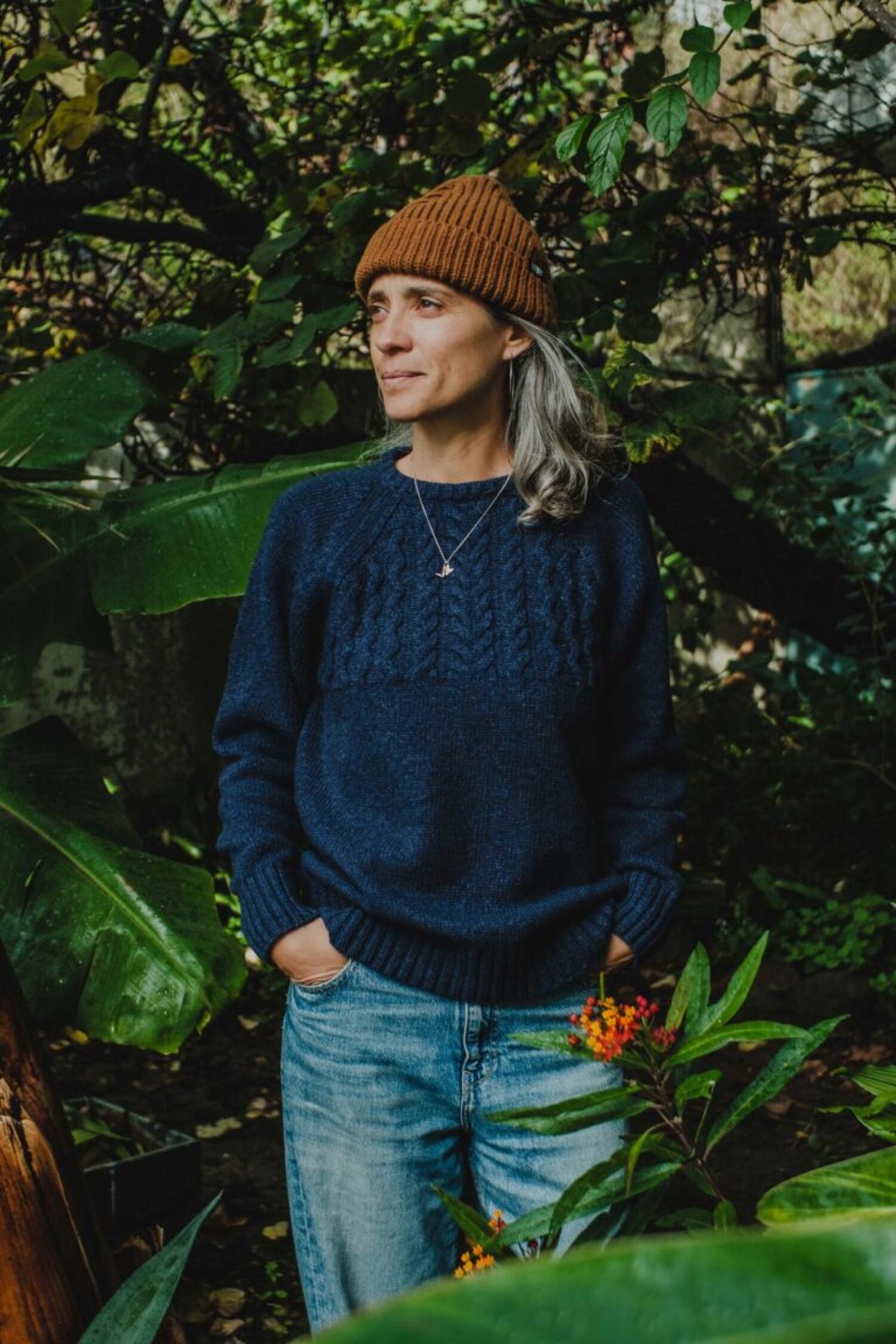 Woman in a lush garden wearing the Lobo Blue Fisherman wool sweater in denim blue and a brown beanie.