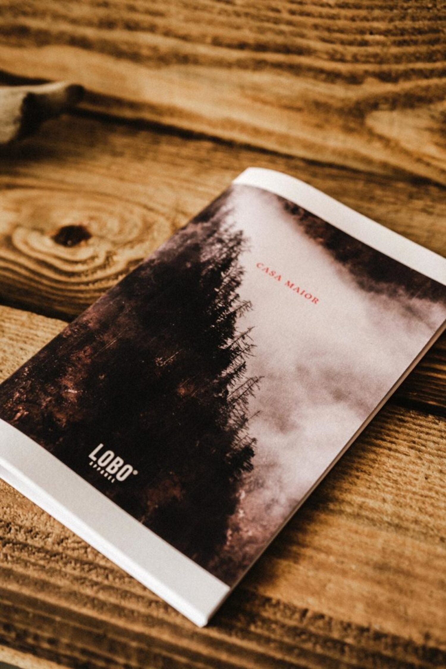 Lobo 'CASA MAIOR' book resting on a rustic wooden table, highlighting the forest photography cover and the Lobo Apparel logo.