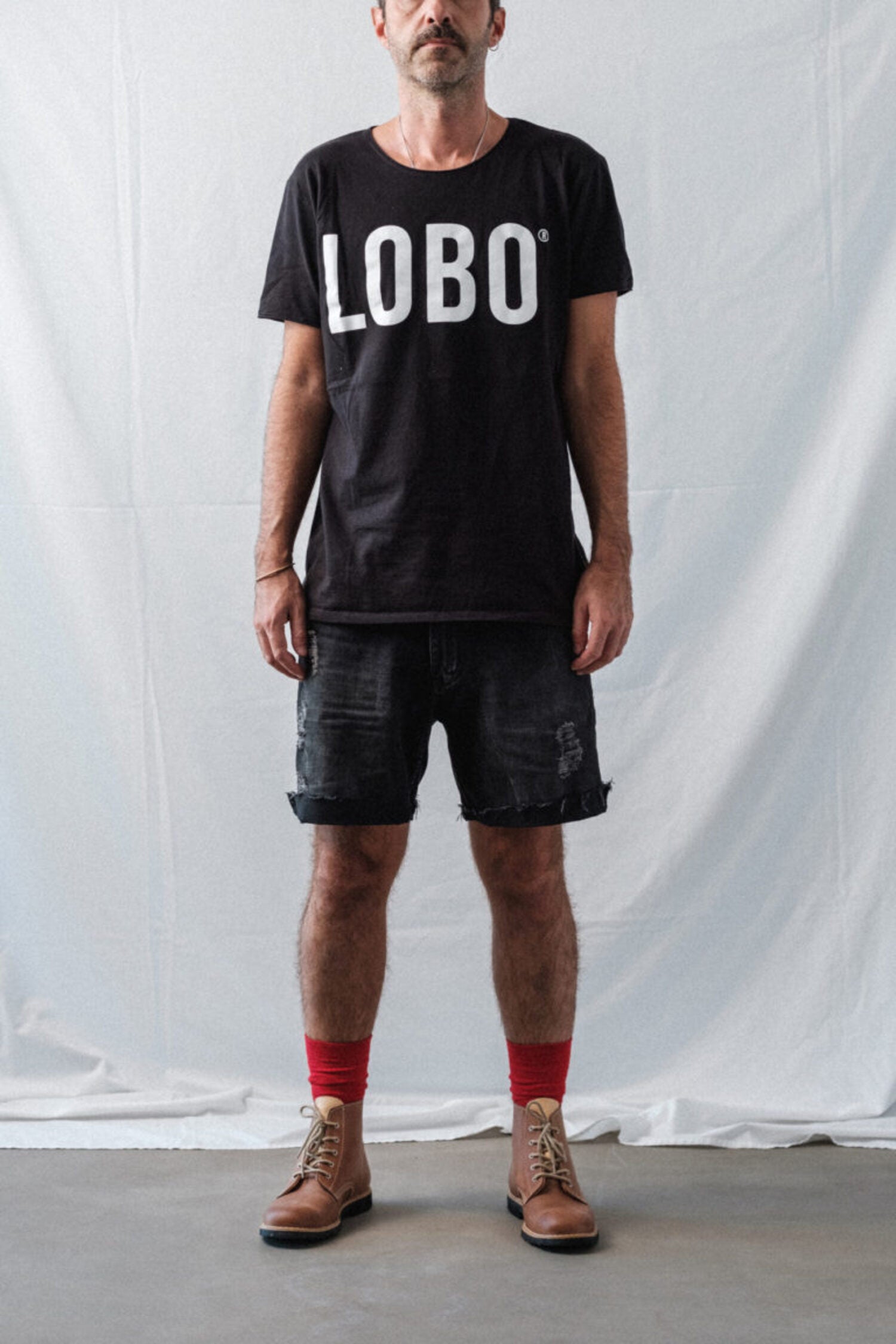 Man wearing a black crew neck t-shirt with Lobo logo, black denim shorts, high red socks, and brown leather boots.